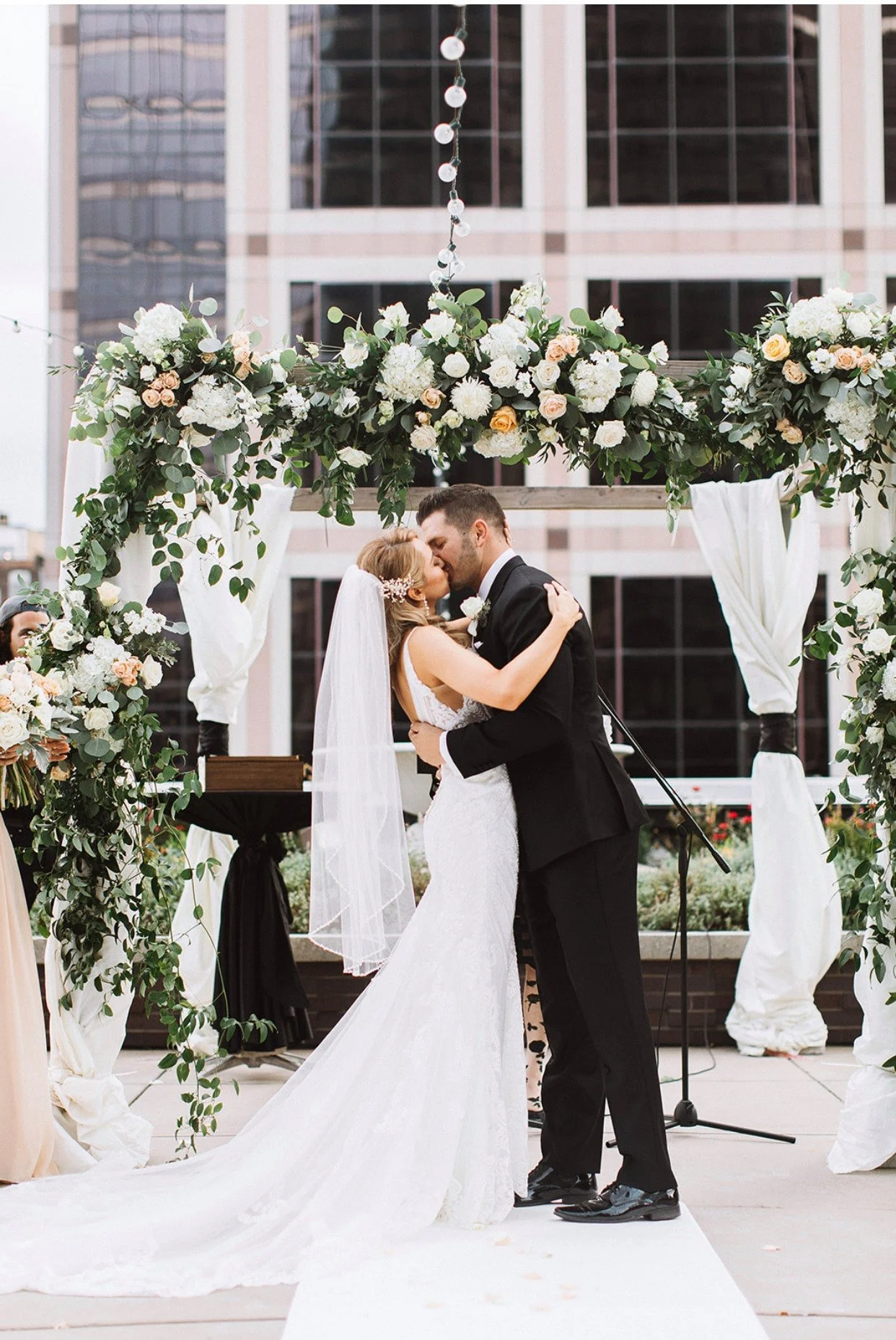 A bride and groom share a kiss during their outdoor wedding ceremony, standing under a floral arch with white and peach flowers and green foliage.
