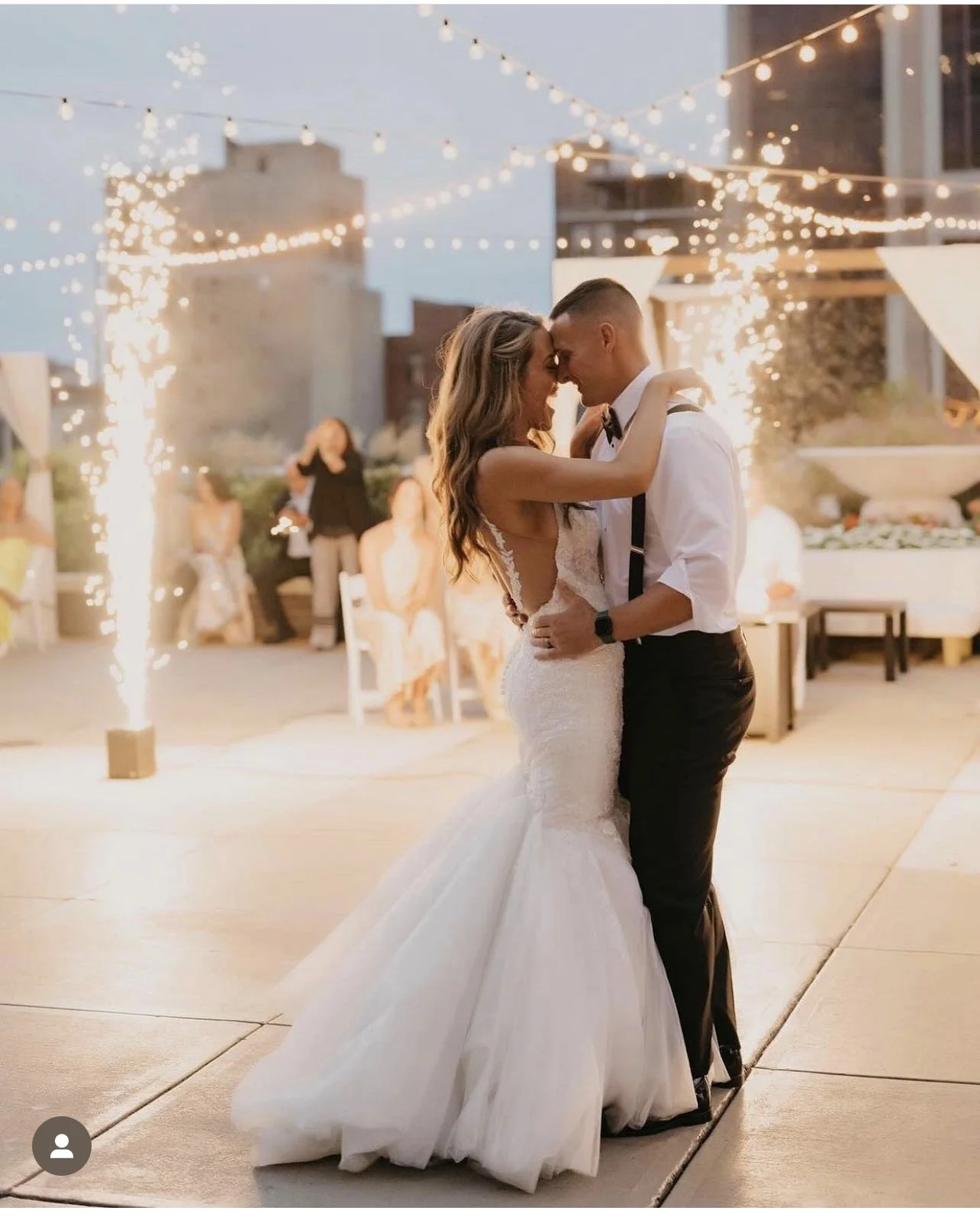 A bride and groom dance at their wedding reception during evening with string lights and sparklers in the background.
