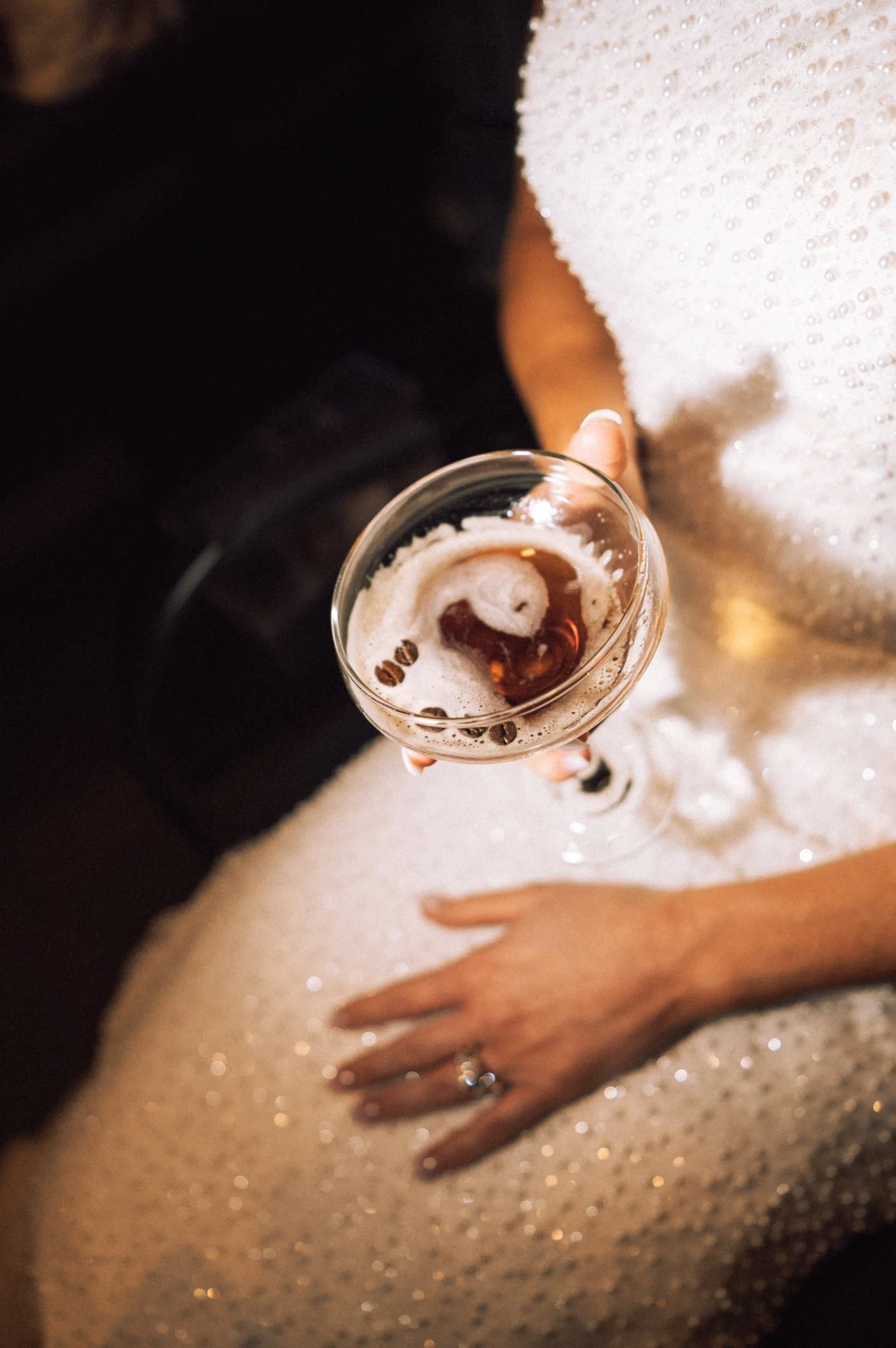 A person in a white dress holding a champagne glass with ice cubes and coffee beans, standing on a sandy beach with a hand resting on their thigh.