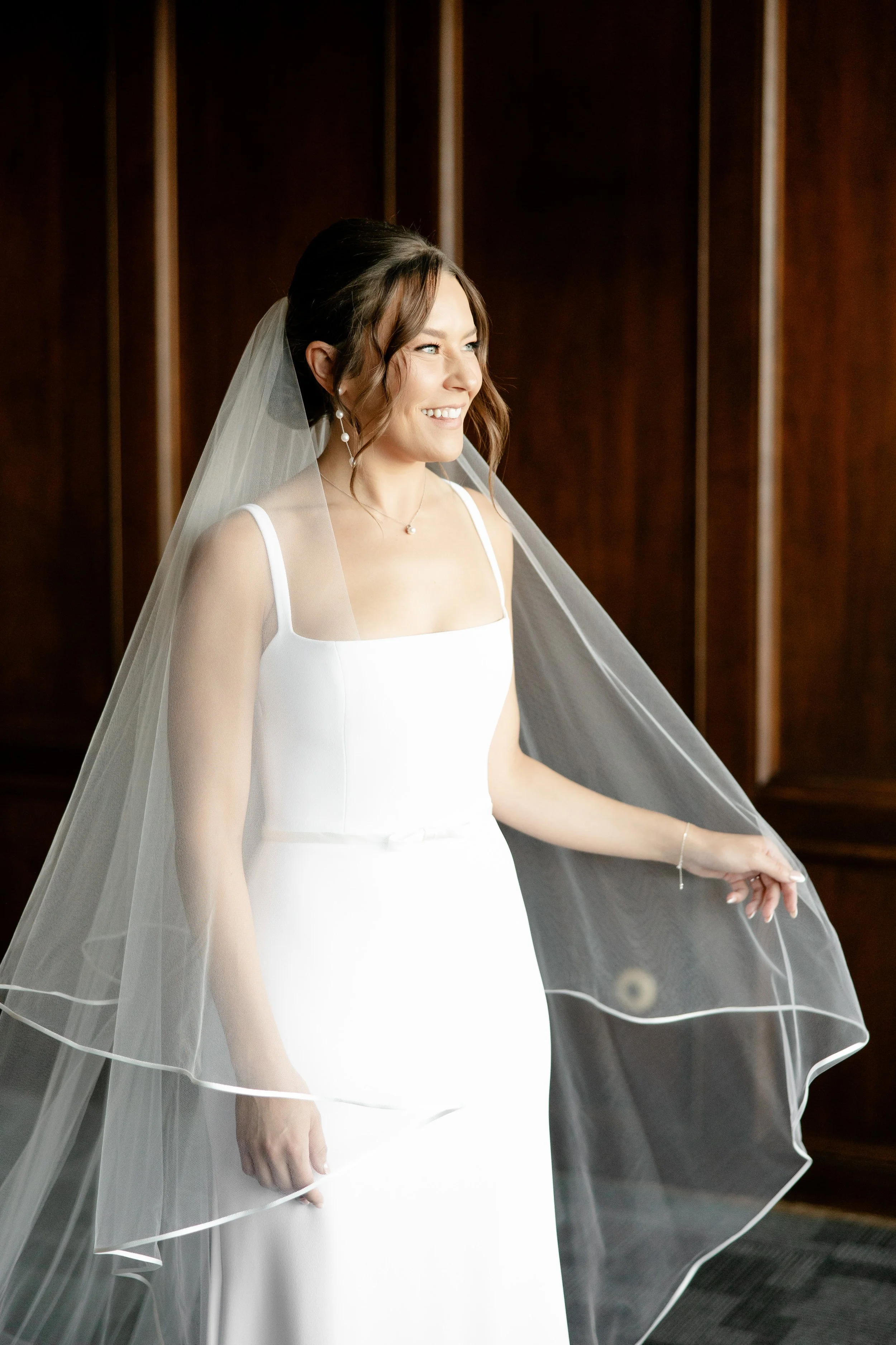 A woman dressed in a white wedding gown with a veil, smiling and looking to her right, standing in front of a dark wooden wall.