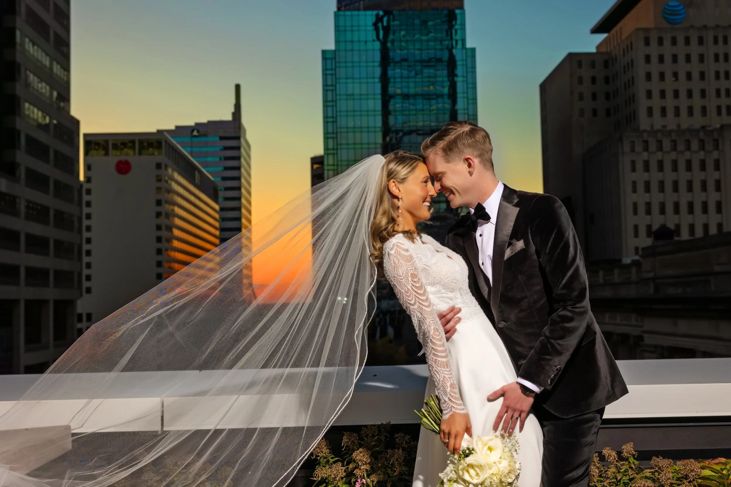 A newlywed couple is standing closely together with their foreheads touching, smiling, and holding a bouquet of white flowers. The bride is wearing a white lace wedding dress and a veil, and the groom is dressed in a black tuxedo with a bow tie, in an urban setting with tall buildings and a sunset in the background.