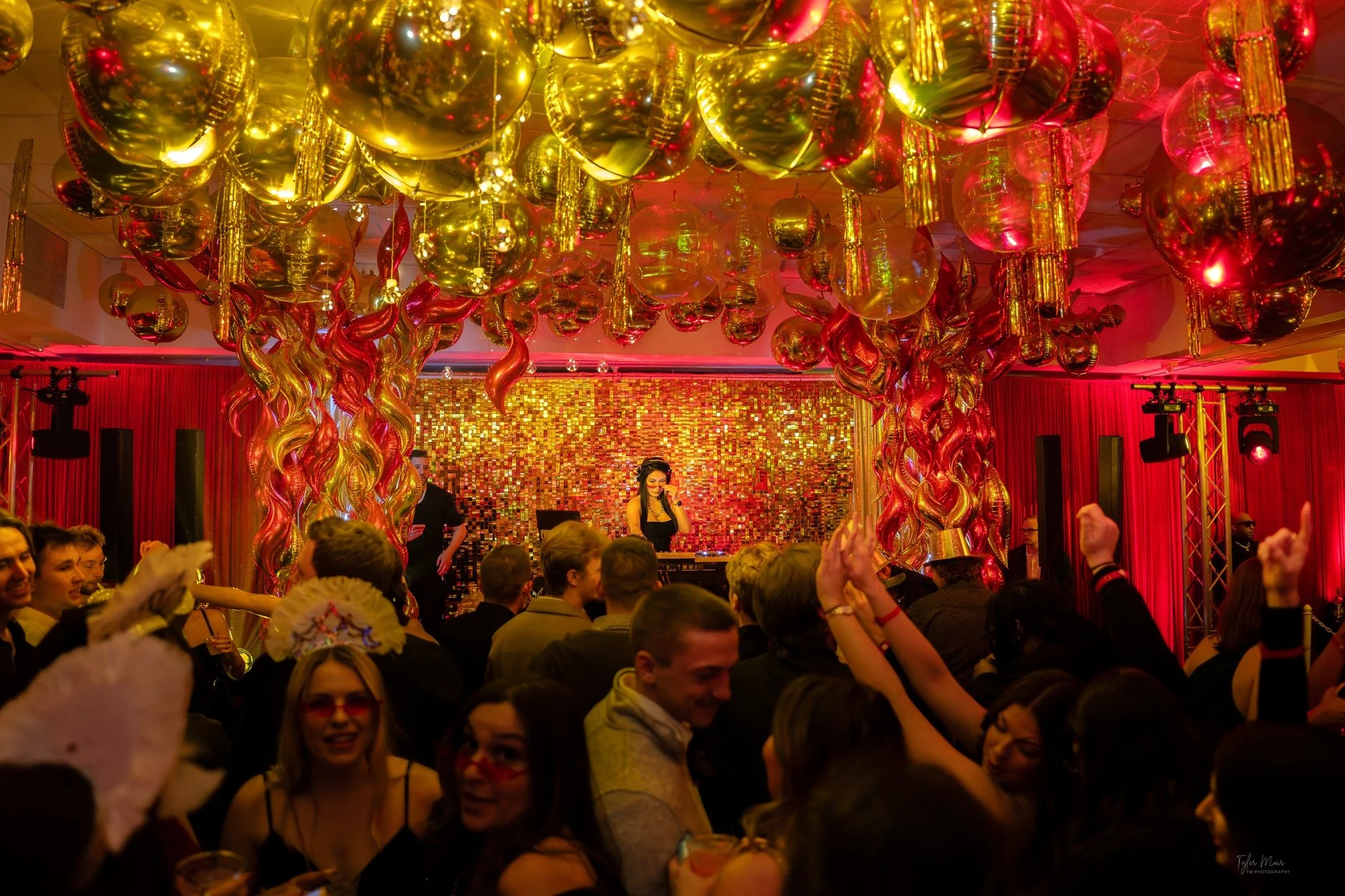 A crowded dance party or celebration with golden and red balloons hanging from the ceiling, a DJ playing music on a stage, and people enjoying and dancing in a lively atmosphere with colorful lighting.