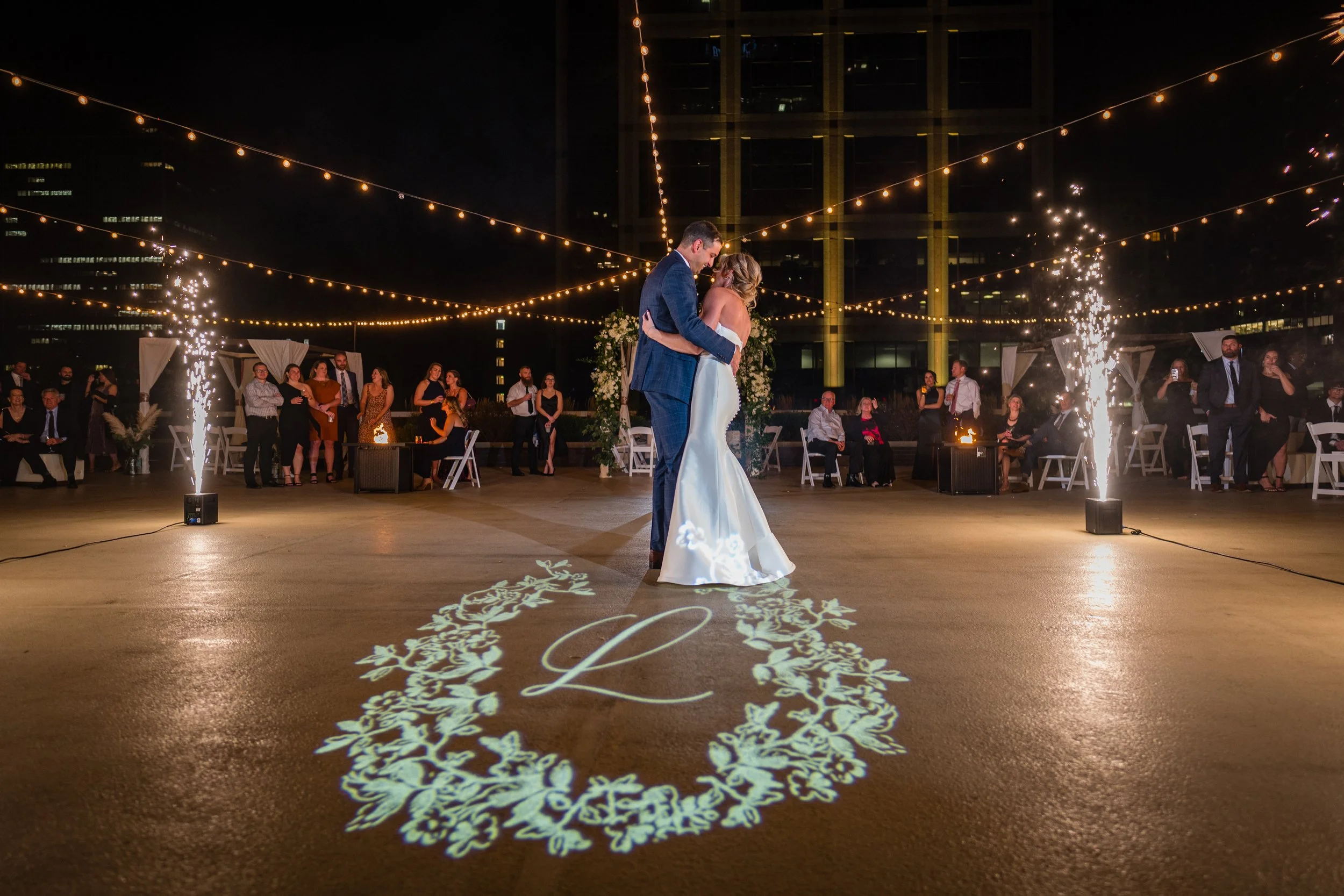 A bride and groom are dancing at their nighttime wedding reception, with guest guests watching and fireworks going off in the background. String lights illuminate the outdoor venue.