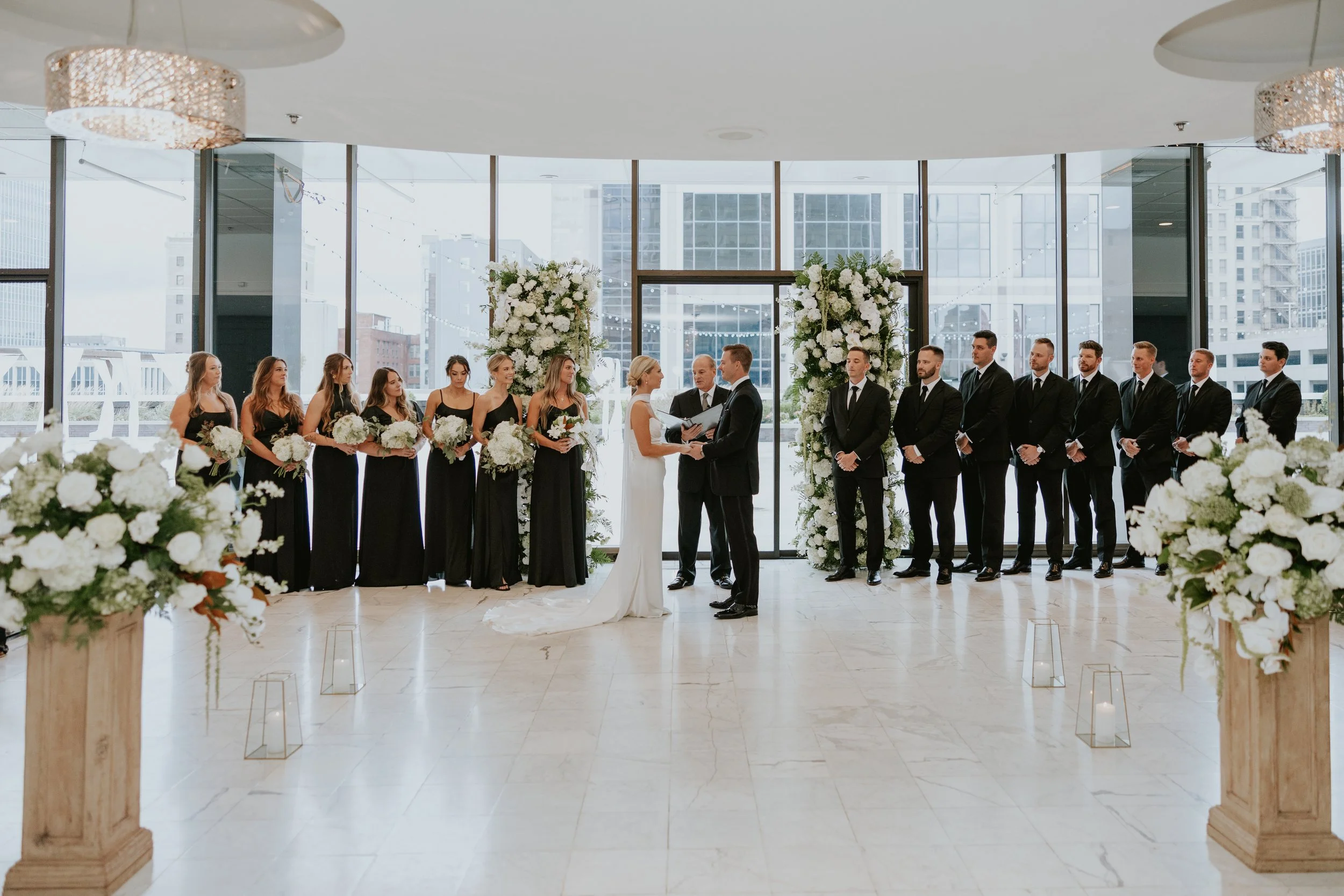 Indoor wedding ceremony in the modern ballroom at JPS Events at Regions Tower, featuring dark wood paneling, brass accents, and custom white floral installations designed by our in-house florists at wholesale pricing