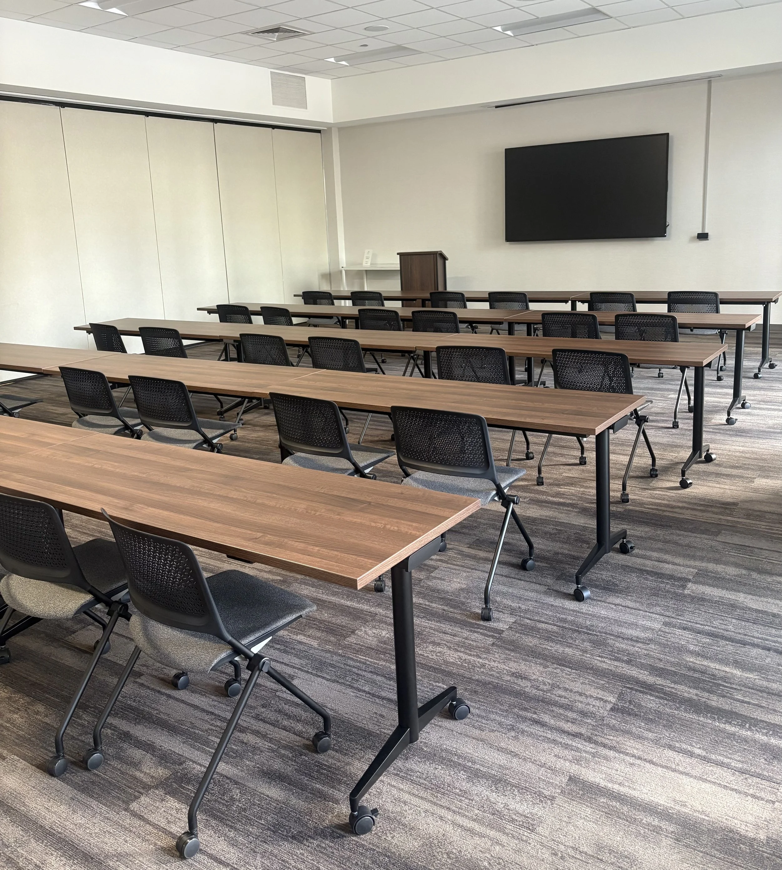 Empty classroom with multiple rows of wooden tables and black mesh chairs on wheels, a large flat-screen TV mounted on the white wall, a small wooden podium, and a white table with a sign or small electronic device. The room has a ceiling with recessed lighting and a gray patterned carpet.