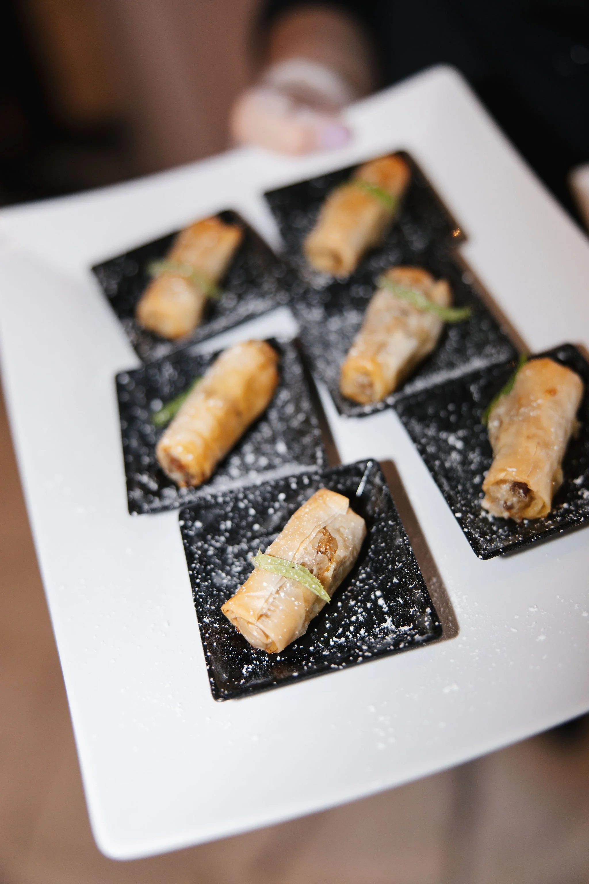 A white rectangular plate with six black squares, each holding a spring roll garnished with a green onion, and lightly dusted with powdered sugar.