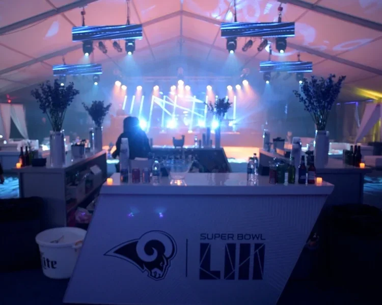 Indoor event space with stage and performance lighting, decorated with white vases filled with tall purple flowers, candles, and beverage bottles. The space features a large tent ceiling with multiple hanging lights, and a front table with the Super Bowl LVI logo, indicating a Super Bowl party or celebration.