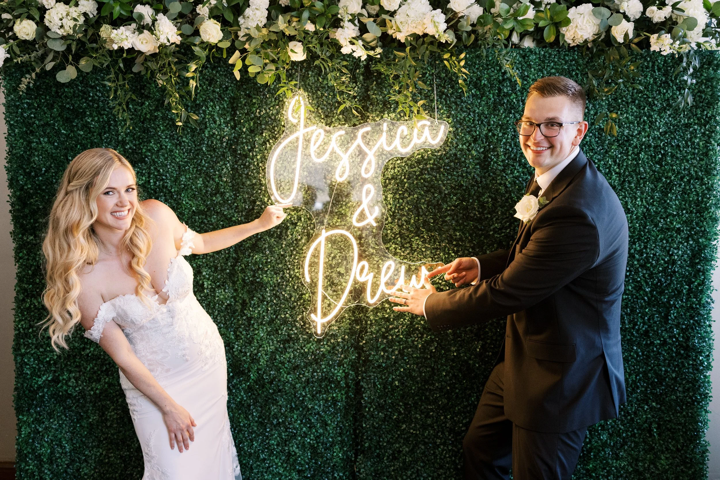 Newlyweds with a custom 'Jessica & Derek' neon sign on a foliage wall at JPS Events at Regions Tower. Highlights in-house floral wall design at wholesale pricing & bespoke photo activations for luxury weddings in downtown Indy.