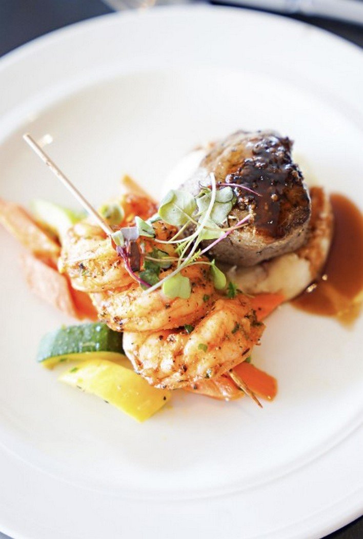 Grilled shrimp and a meat ball with sauce, garnished with microgreens, served on a bed of vegetables and mashed potatoes.