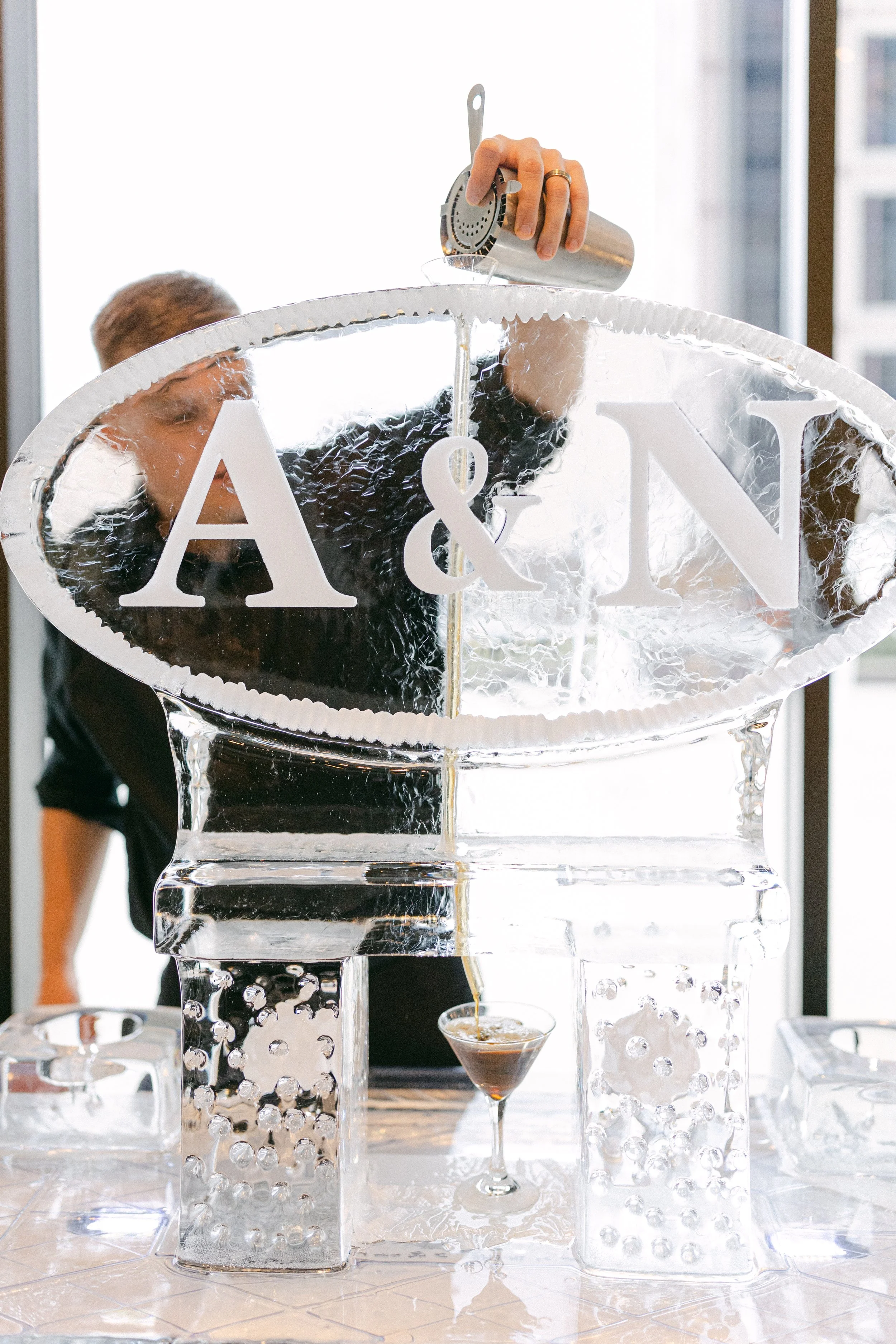 A person pours coffee from a metallic coffee maker into a glass below. The setup includes clear ice sculptures and glassware with coffee drinks. The scene is indoors with large windows in the background.