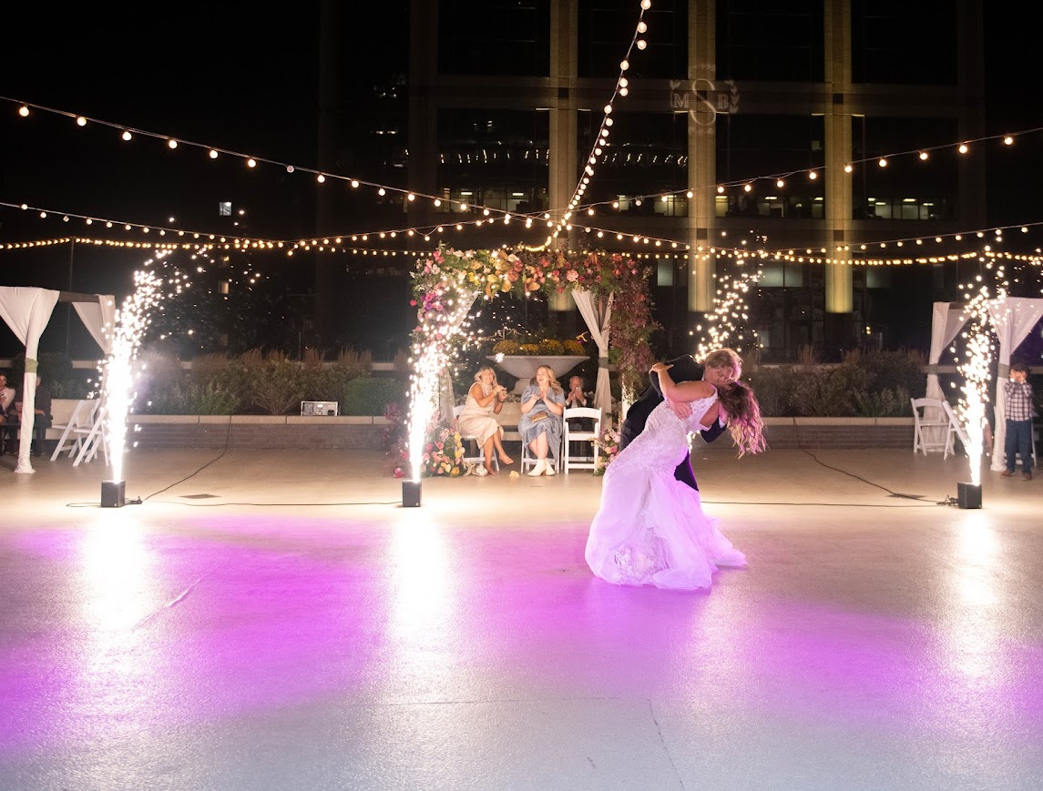 A bride and groom dance at their wedding reception with sparklers and string lights in the background at night.