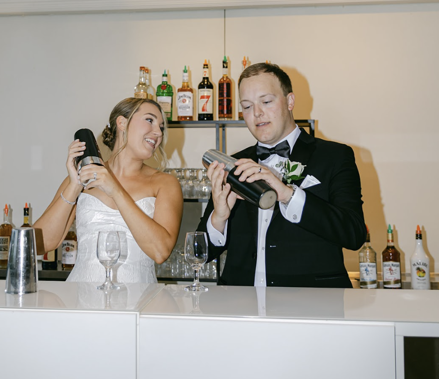 Bride and groom at a bar, smiling, with bottles of alcohol displayed behind them, as they prepare drinks.