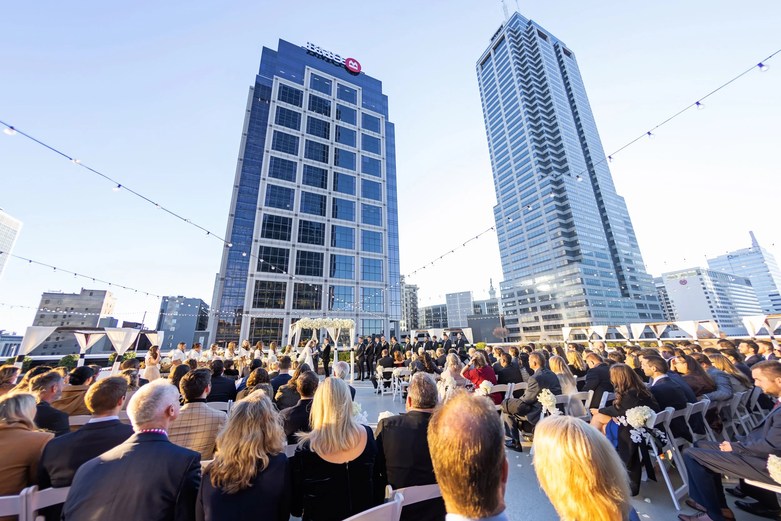 People attending a wedding ceremony on a rooftop with city skyscrapers in the background, under string lights on a clear day.