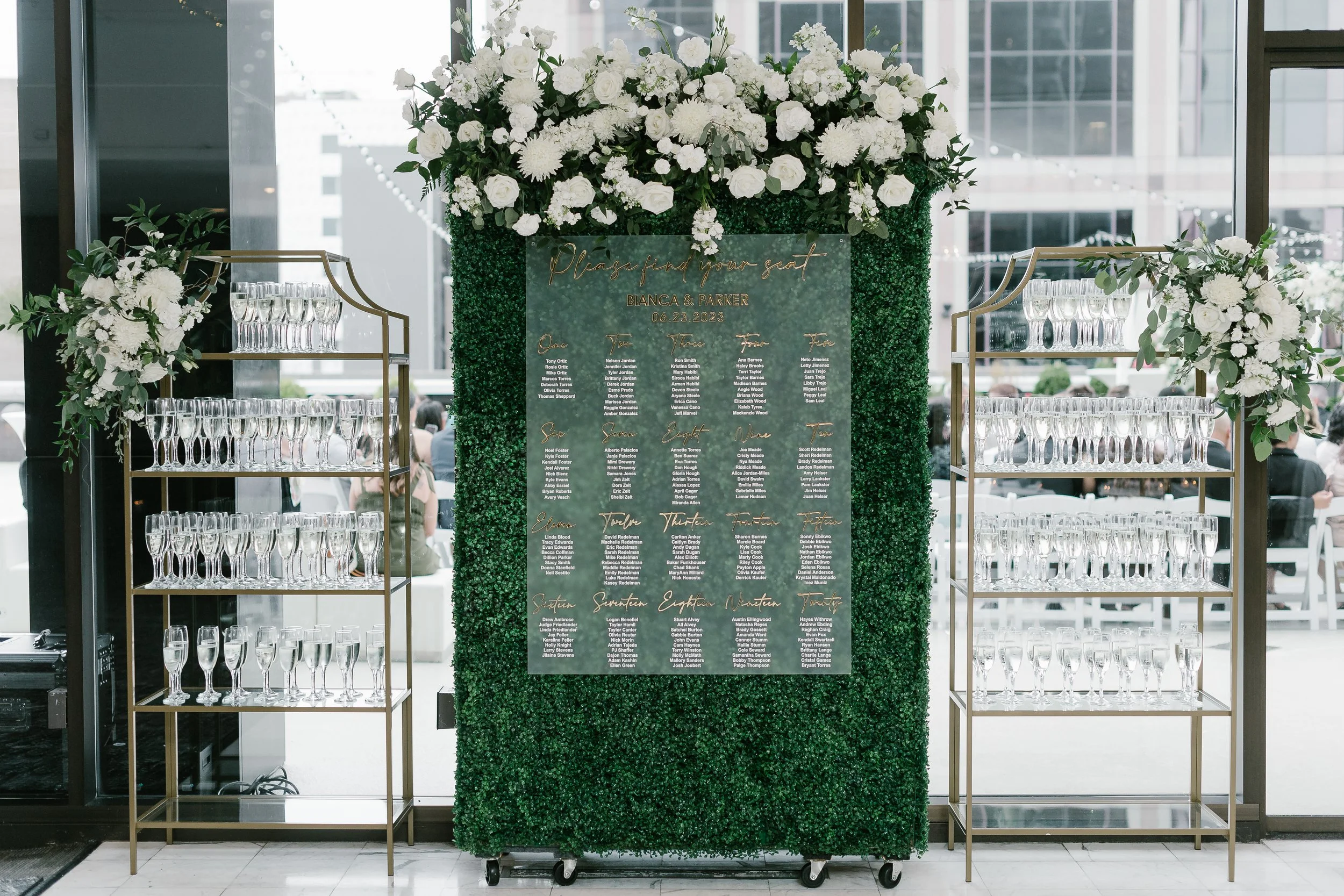 A large wedding seating chart with white flowers and greenery, flanked by two gold shelving units filled with empty champagne glasses, positioned in a modern building with glass windows.