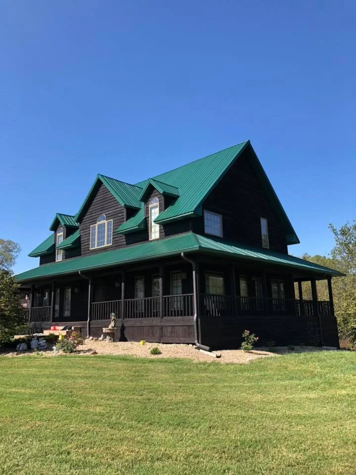 A large, black, two-story house with a green metal roof and a spacious front porch.