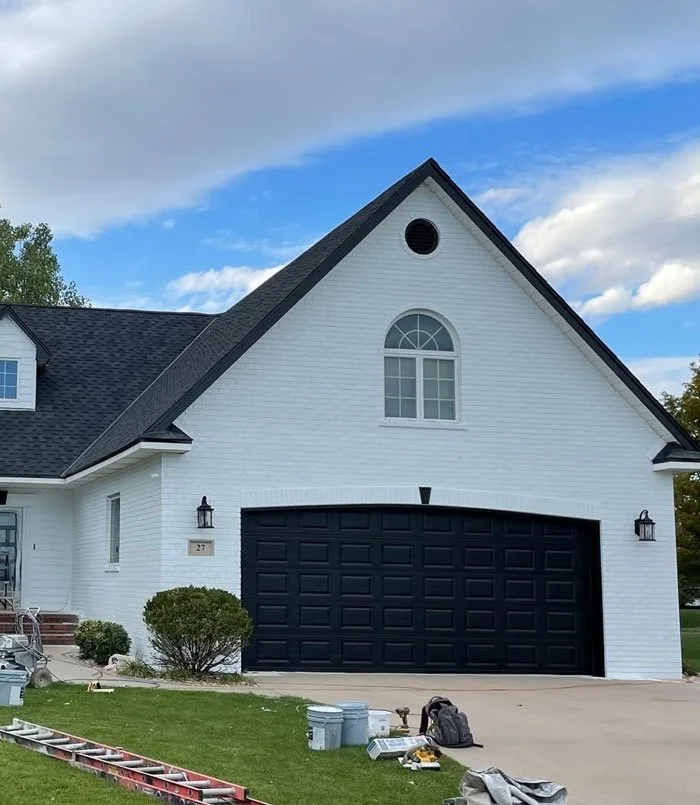 A white house with a black garage door, black lantern-style exterior lights, a small front tree, and a dormer window on a sunny day with some clouds in the sky.