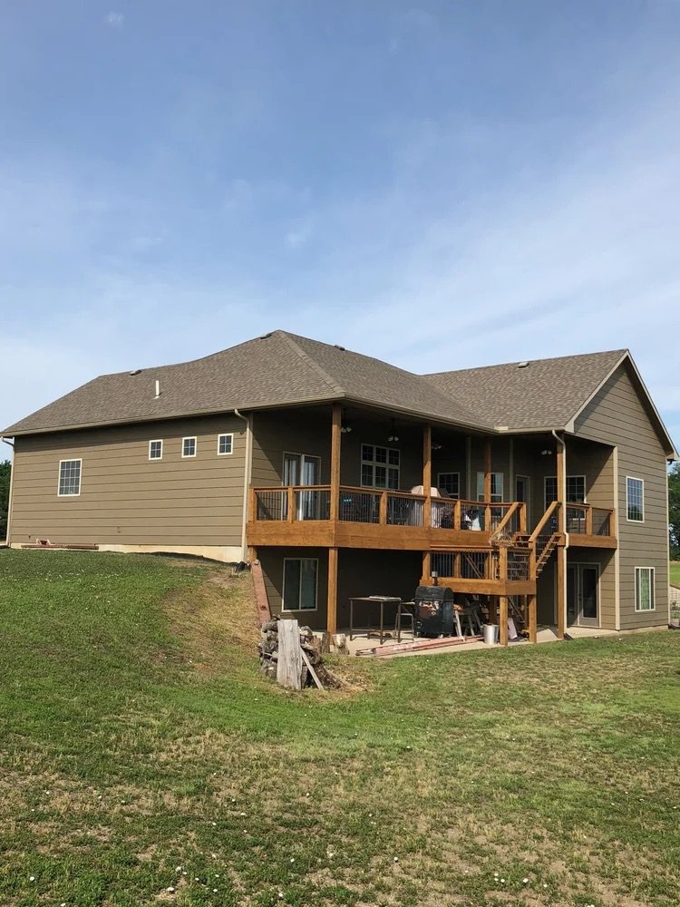 A two-story house with a newly constructed wooden deck on the back, overlooking a grassy yard under a clear blue sky.