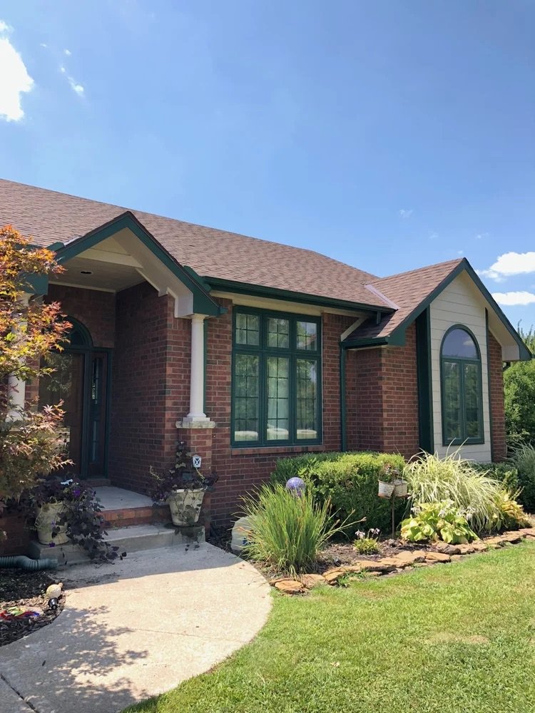 Front view of a brick house with large green-trimmed windows, a concrete pathway, and a landscaped yard with various plants and shrubs.