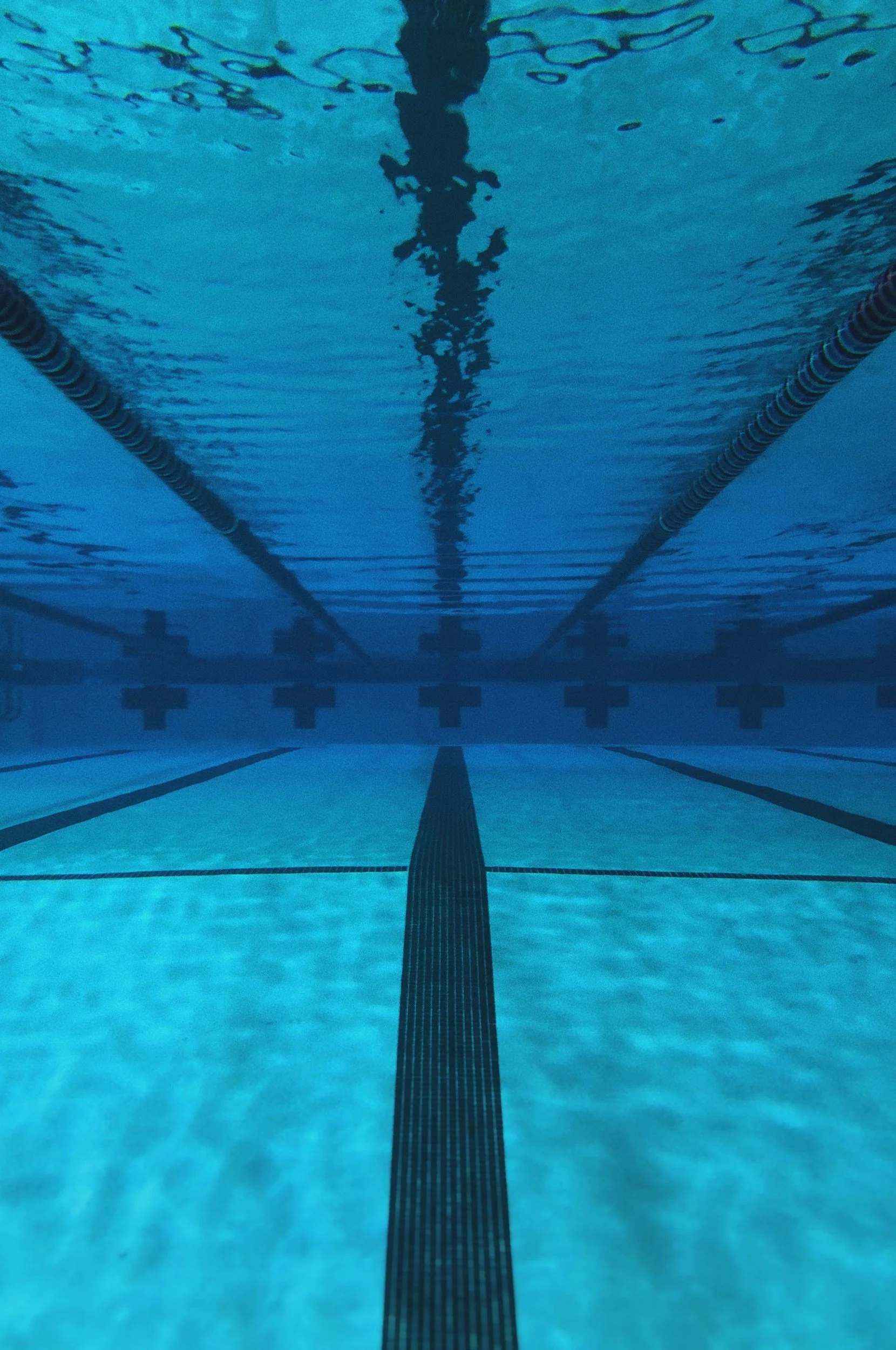 Underwater view of a swimming pool with lane markers and a swimmer's reflection on the water surface.