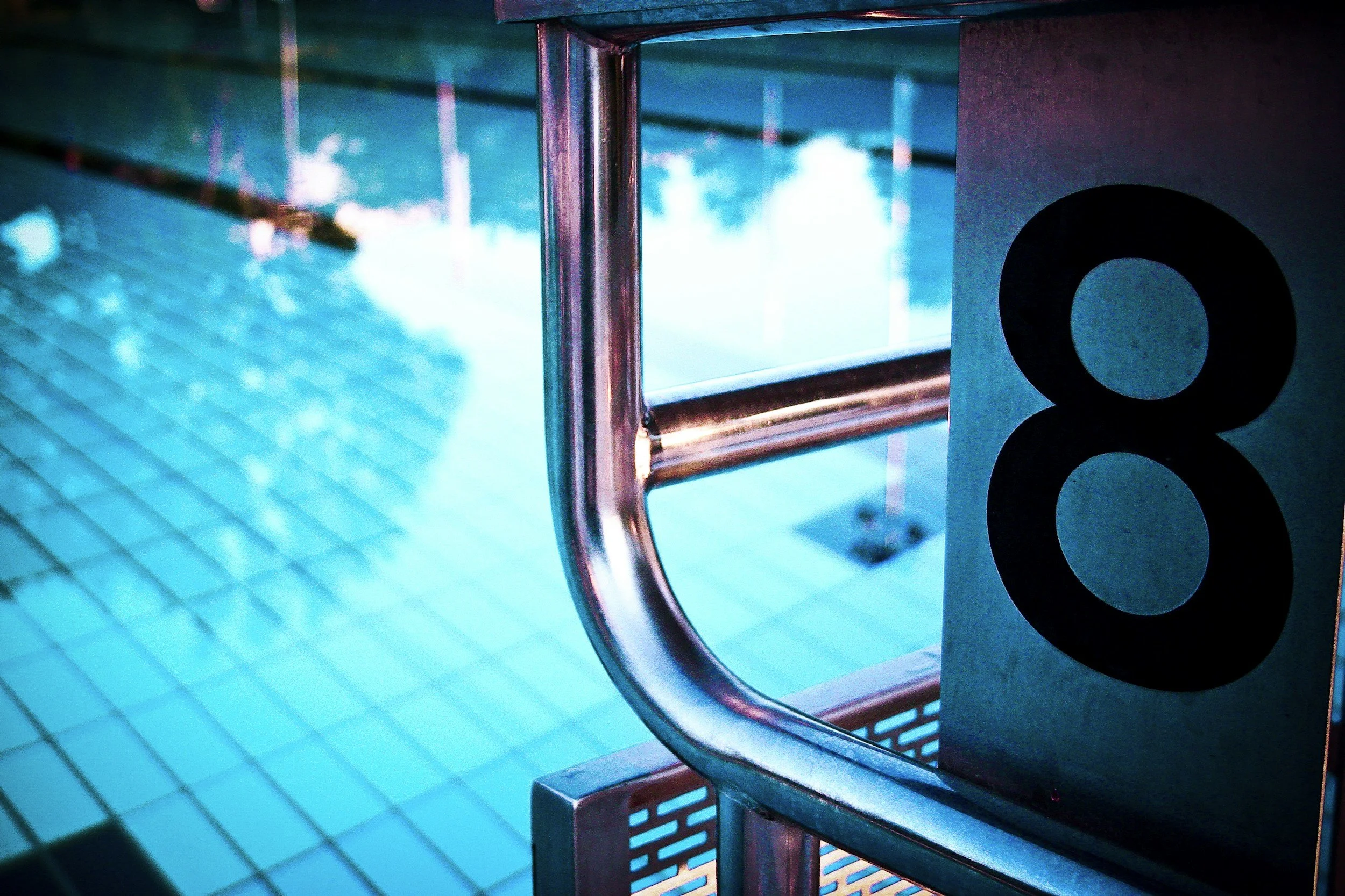 Close-up of a stainless steel ladder in a swimming pool, with a numbered sign in the foreground reading '8', against blue pool tiles and clear water.