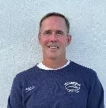 A man with short hair wearing a navy blue athletic shirt, standing against a plain, light wall background.