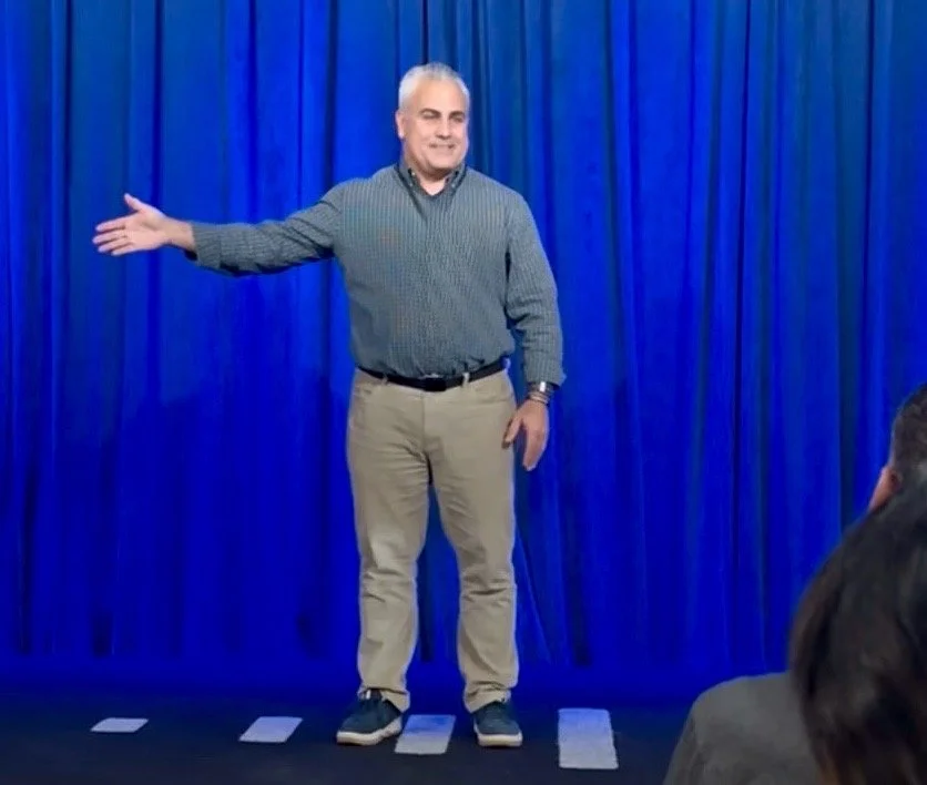 A man with gray hair standing on a stage with blue curtains, gesturing with one arm extended, wearing a checkered shirt and beige pants.