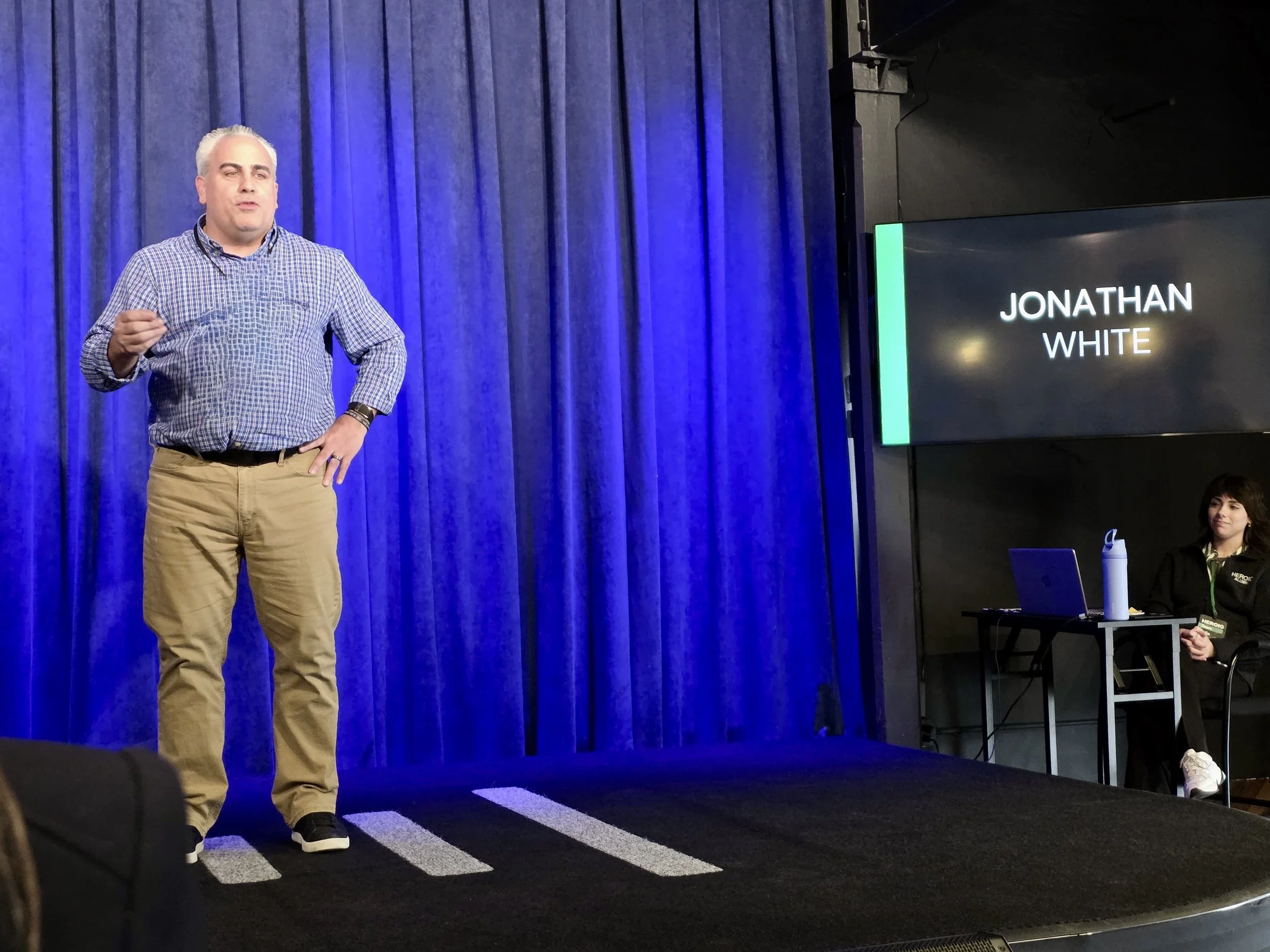A man with gray hair and a checked shirt speaking on stage in front of blue curtains, with a large screen displaying the name "Jonathan White" beside him. A woman is seated at a table with a laptop and water bottle, watching.