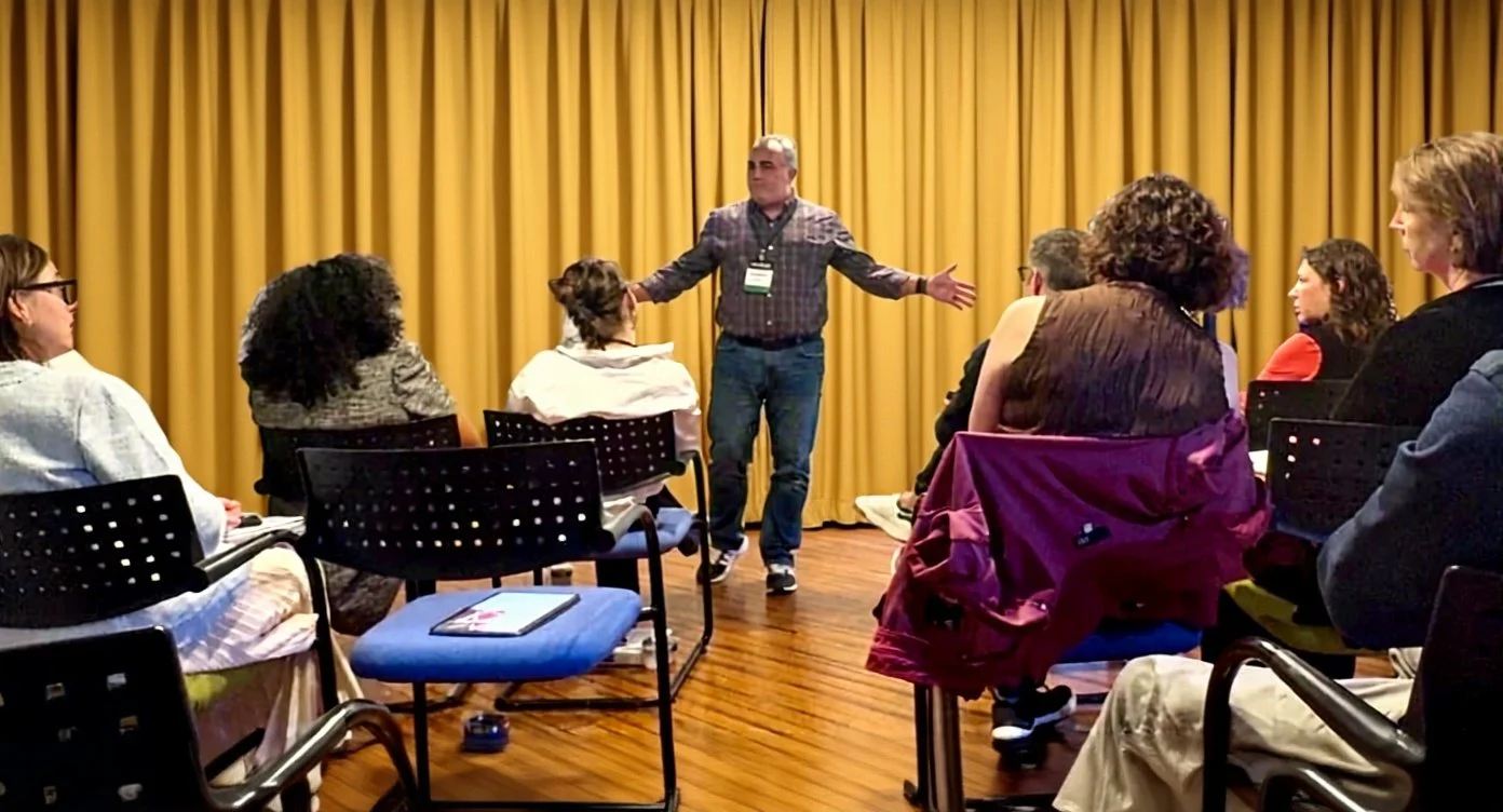 A man giving a presentation in front of a group of seated people in a room with a golden curtain backdrop.
