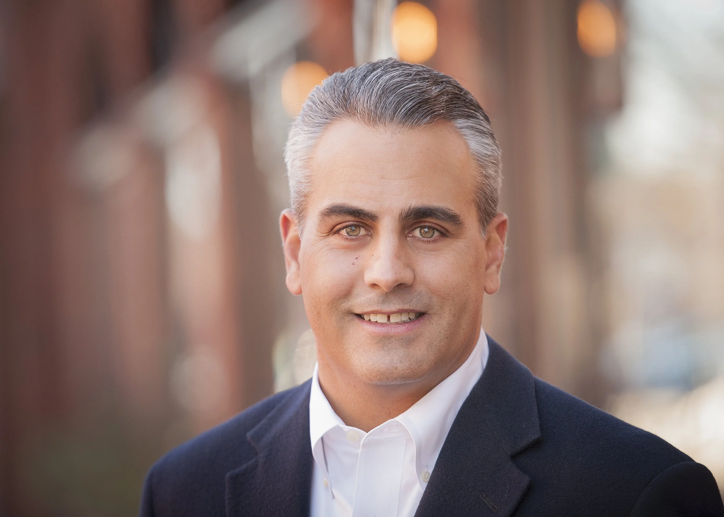 Portrait of a middle-aged man with gray hair, wearing a dark blazer and white shirt, smiling outdoors with blurred background and warm lighting.