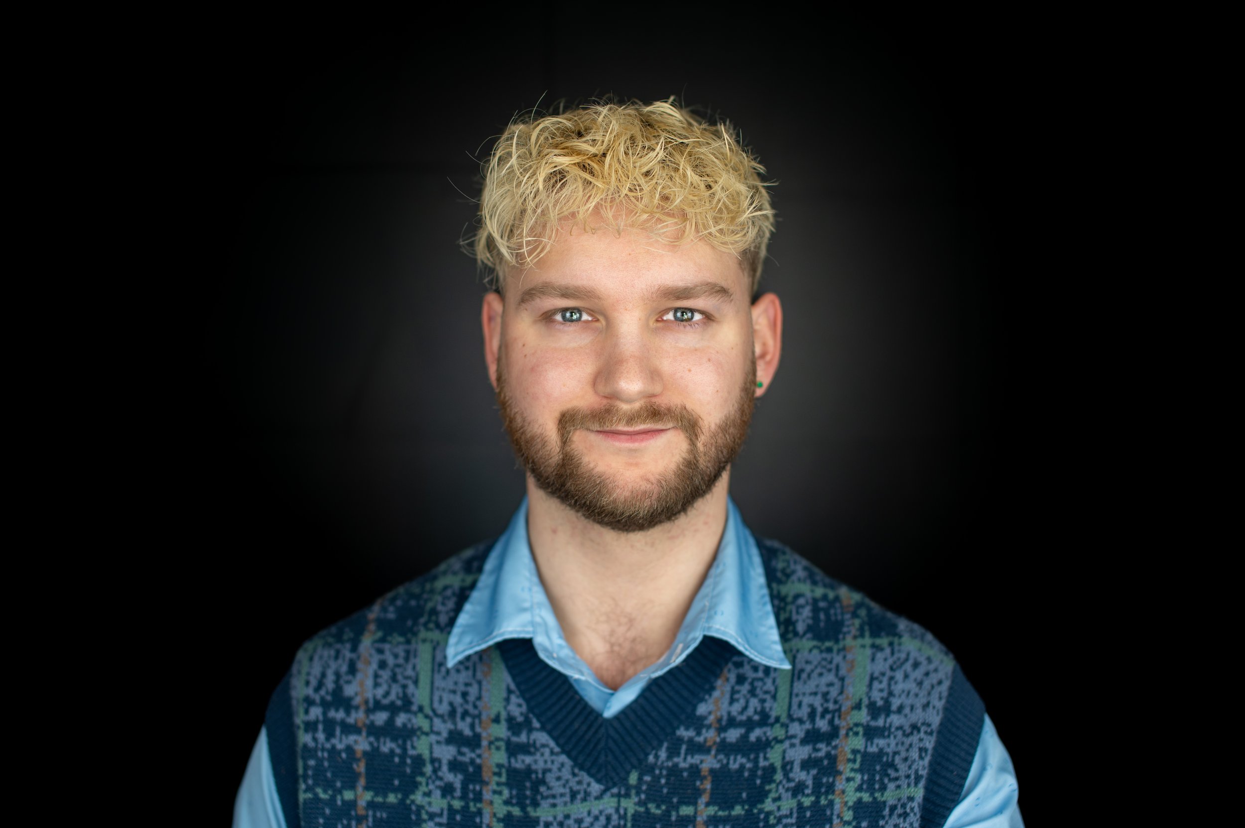 Portrait of a young man with blonde curly hair, blue eyes, and a beard, wearing a blue collared shirt and a patterned sweater vest, against a black background.