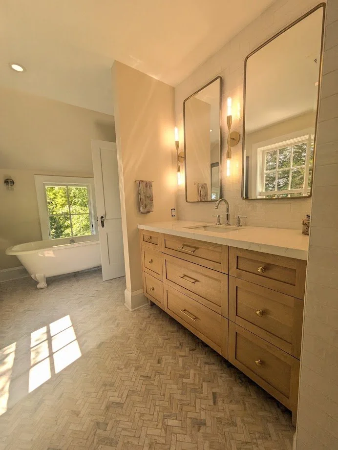 A bathroom featuring a wooden vanity with a white countertop, two large mirrors, modern wall sconces, a window reflecting greenery, a white clawfoot bathtub, and herringbone patterned tile flooring.