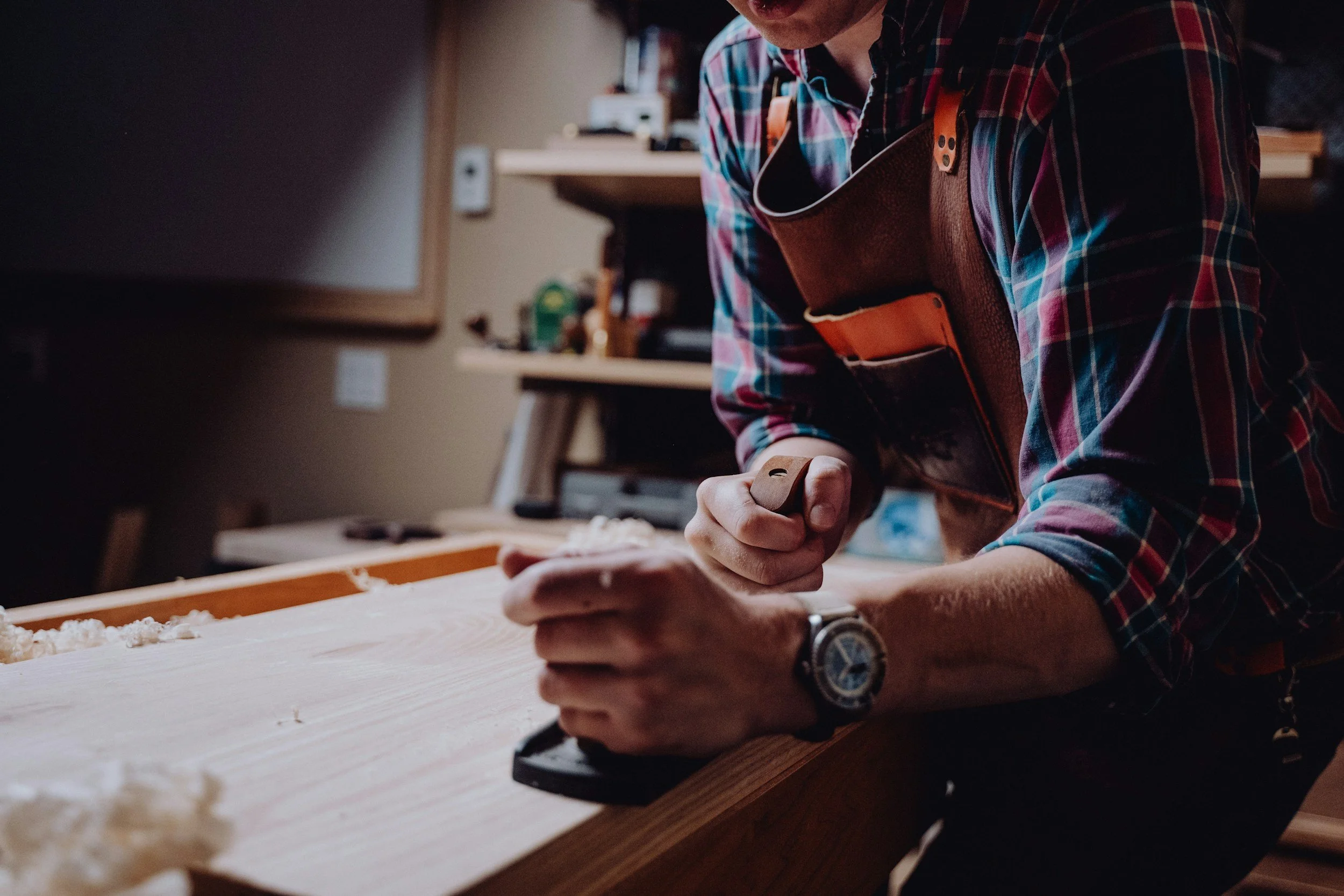 A person in a plaid shirt with an apron is using a hammer and chisel on a piece of wood in a workshop. Shelves with tools and supplies are visible in the background.