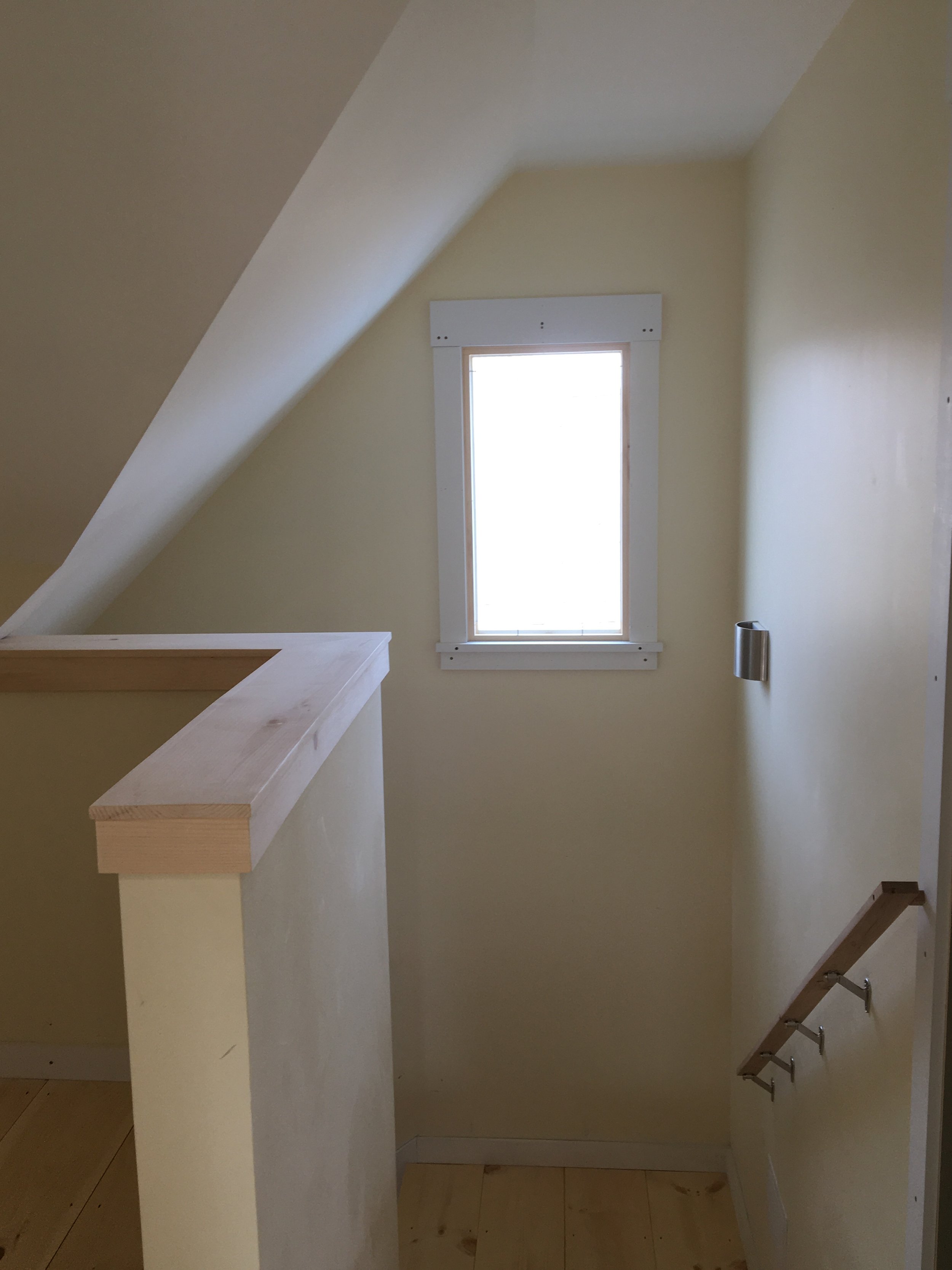 An interior view of a room with a small window, a wooden ledge, a white wall, and a staircase with metal railings.