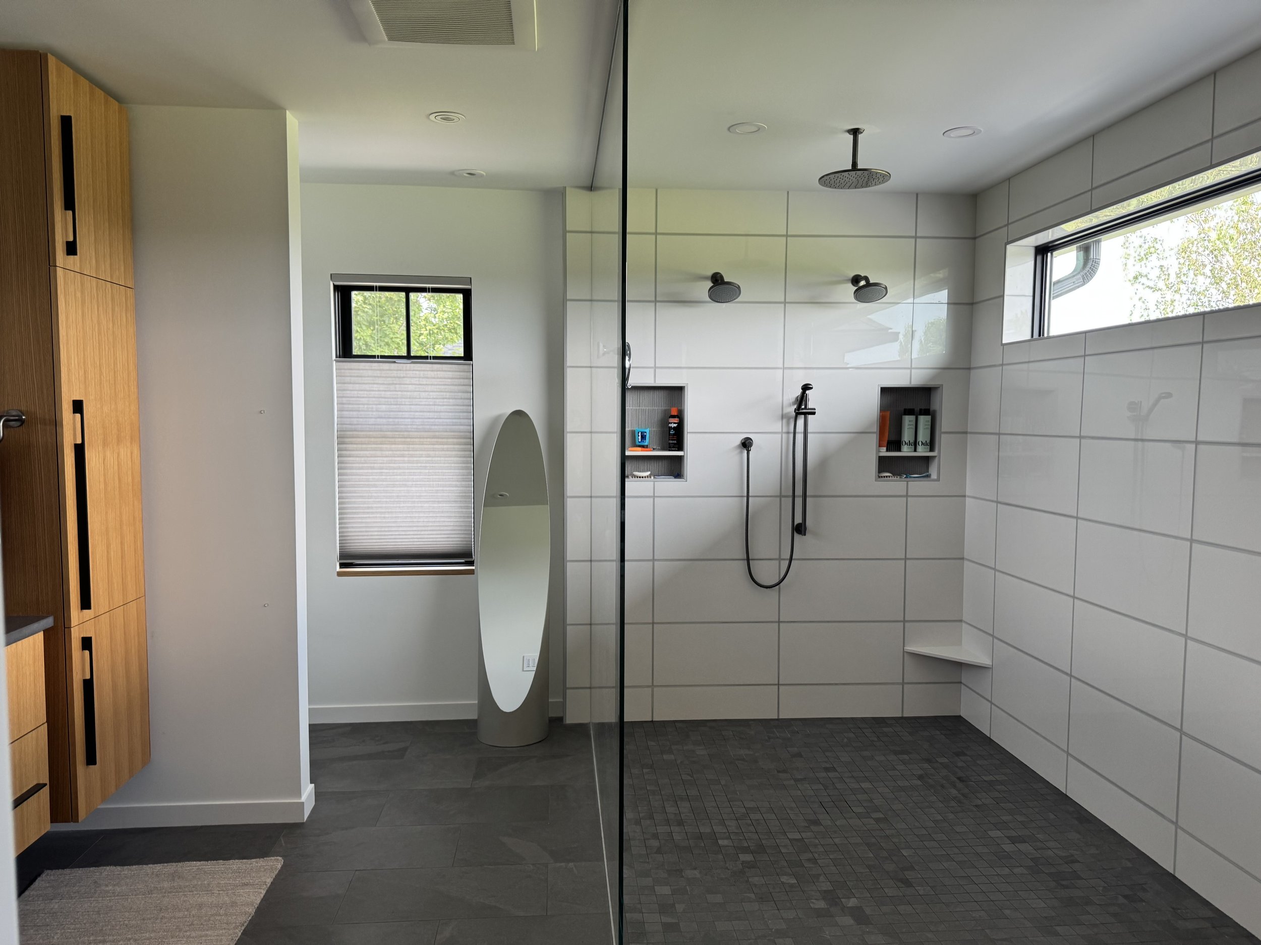 Modern bathroom with a glass shower enclosure, white tiled walls, wooden cabinetry, a window, and a floor mat.