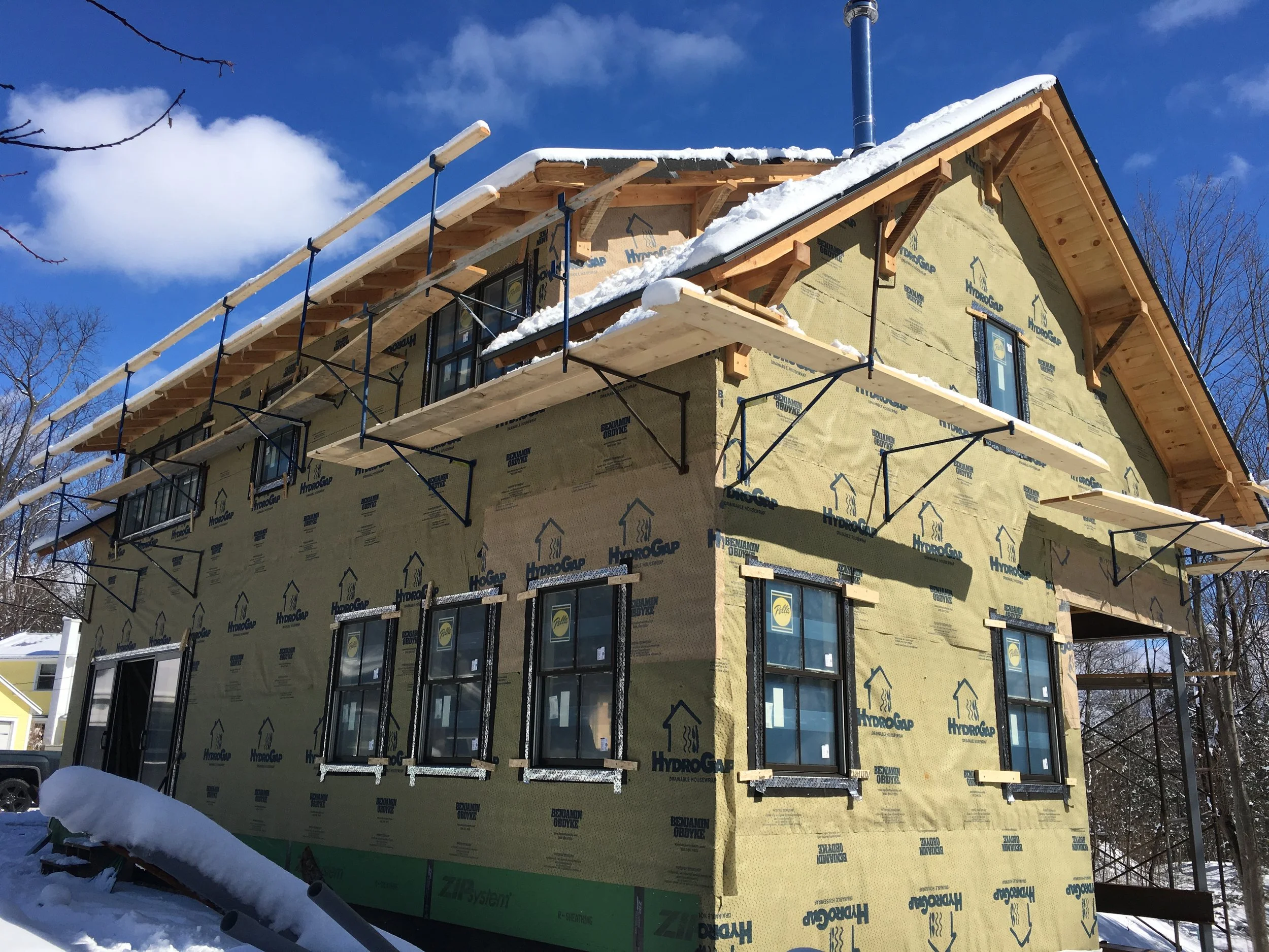 A house under construction with yellow insulation wrapped around the exterior, snow on the roof, and scaffolding around it in a winter setting.