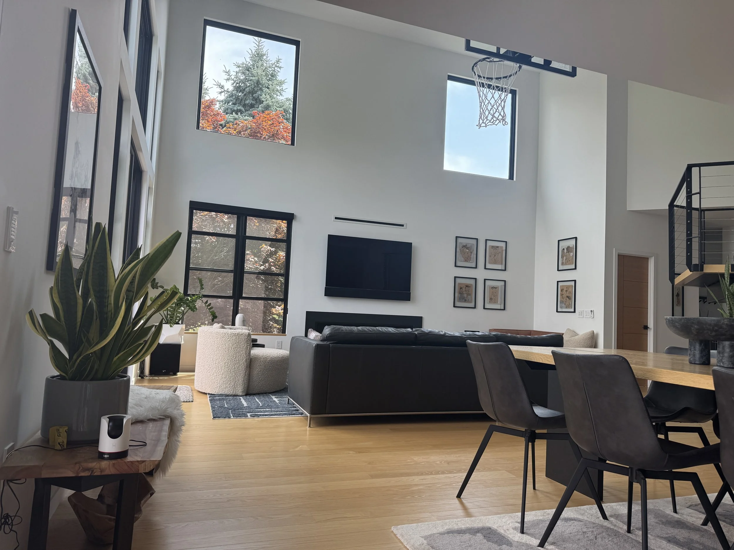 Modern living room with high ceiling, large windows showing trees outside, black and beige furniture, wall art, and green plants.