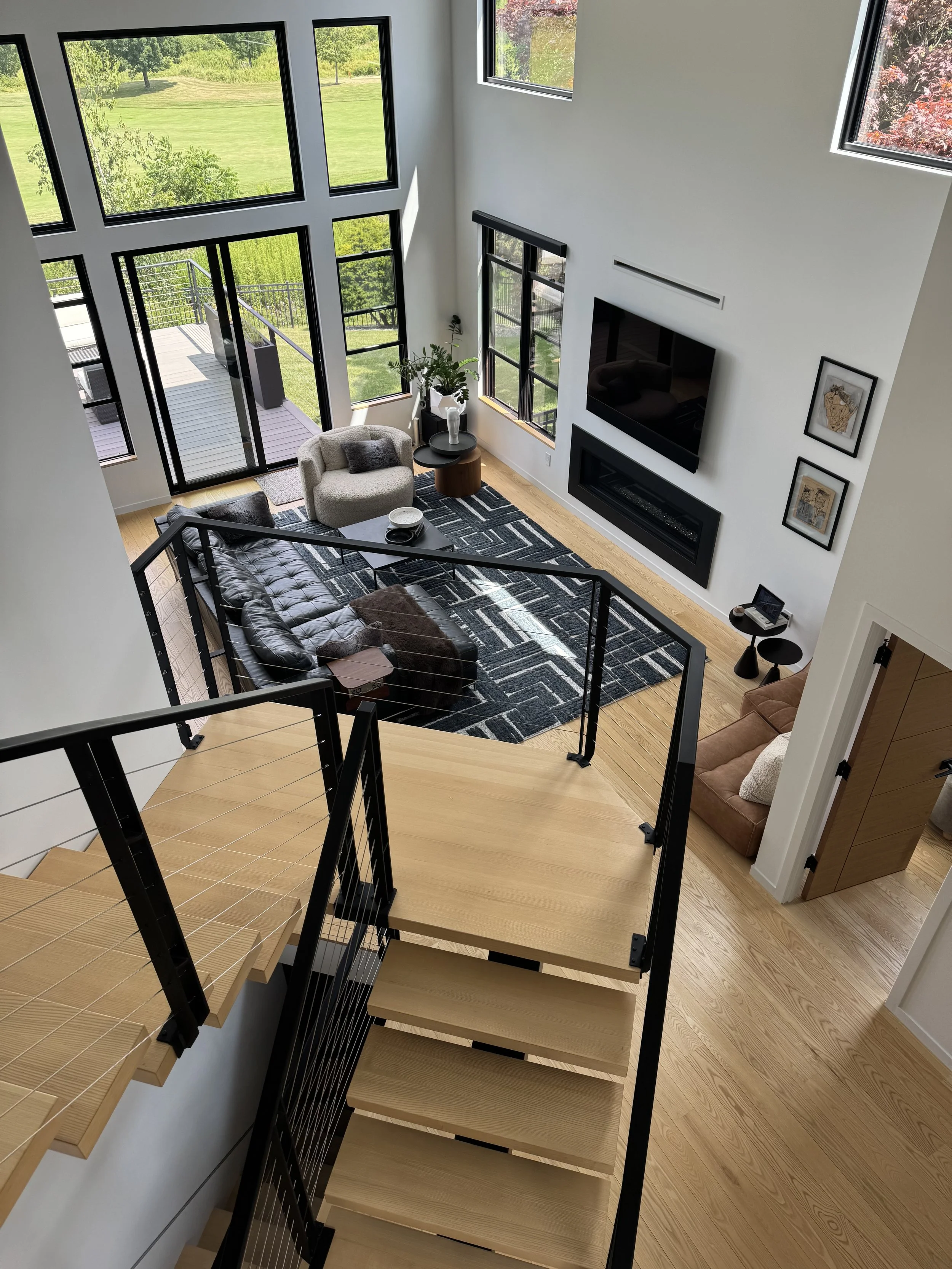 Living room with large windows showing a green outdoor landscape, a black leather sofa with throw pillows, a beige armchair, black and wooden side tables, a patterned rug, a wall-mounted TV, and framed artwork.