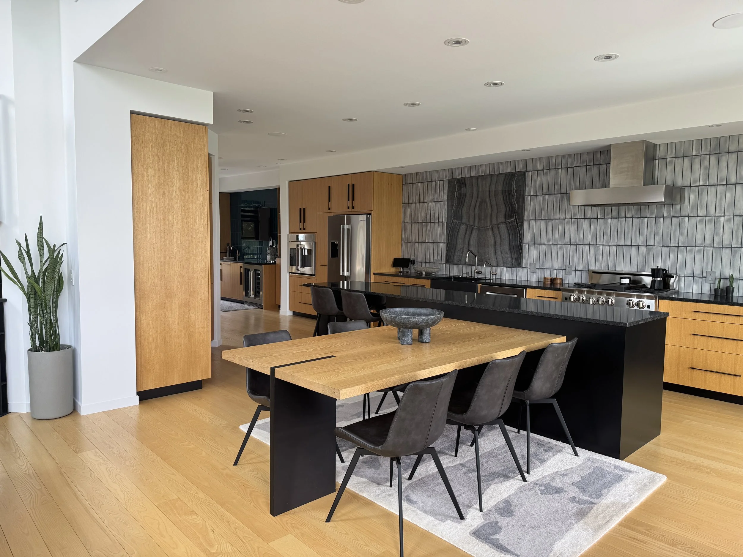 Modern kitchen with black countertop, wooden cabinets, stainless steel appliances, gray tiled backsplash, black chairs around a wooden dining table, gray and white patterned rug, a potted plant near the wall, and recessed ceiling lights.