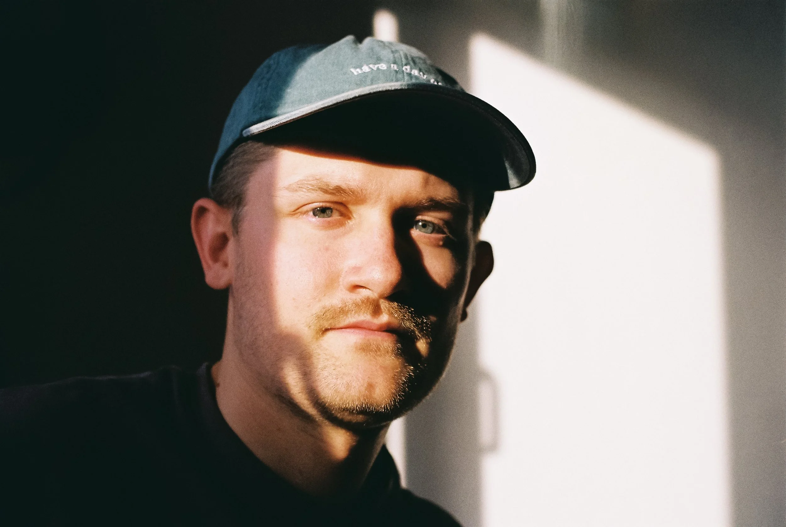 Junger Mann trägt grauen Cap, Blick in die Kamera, Sonnenlicht fällt auf sein Gesicht, dunkler Hintergrund, dunkles T-Shirt.