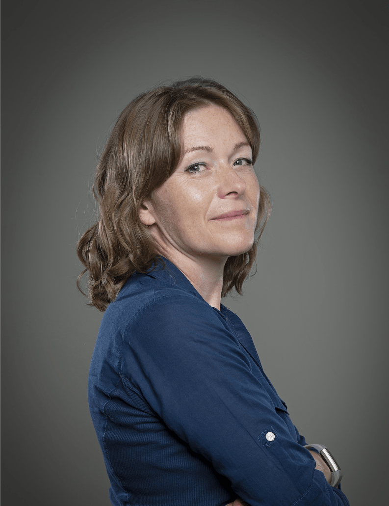 A woman with wavy, shoulder-length brown hair wearing a dark blue shirt, standing with her arms crossed, facing slightly to the right, against a gray background.