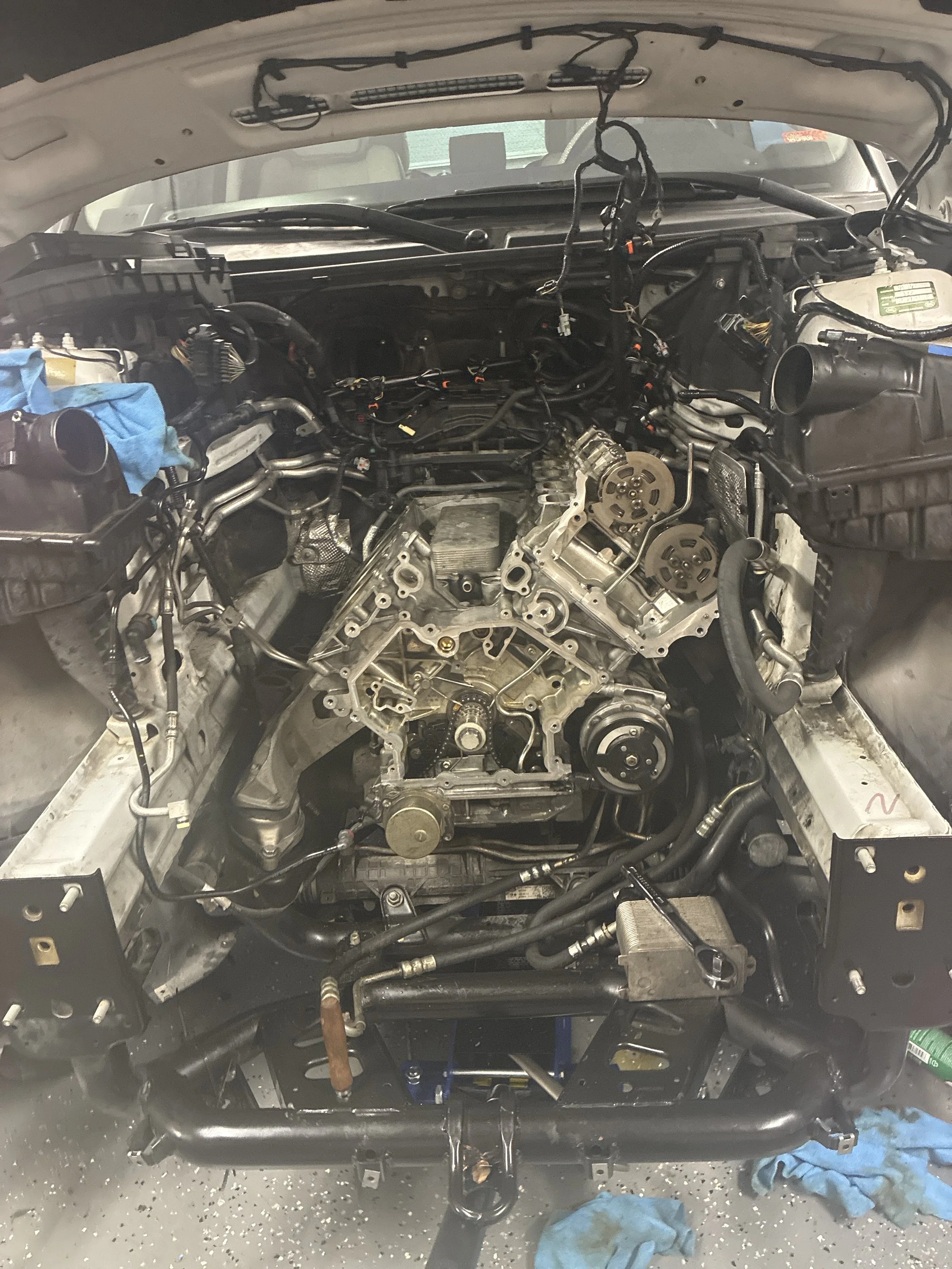 Car engine bay with engine removed, exposing internal components and wiring, with blue shop towels and tools on the ground.