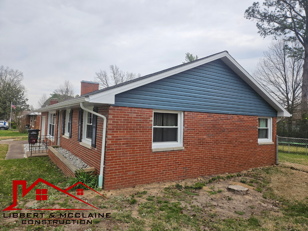 Single-story brick house with blue siding and white trim, front yard with patchy grass, and a chain-link fence on the right side.