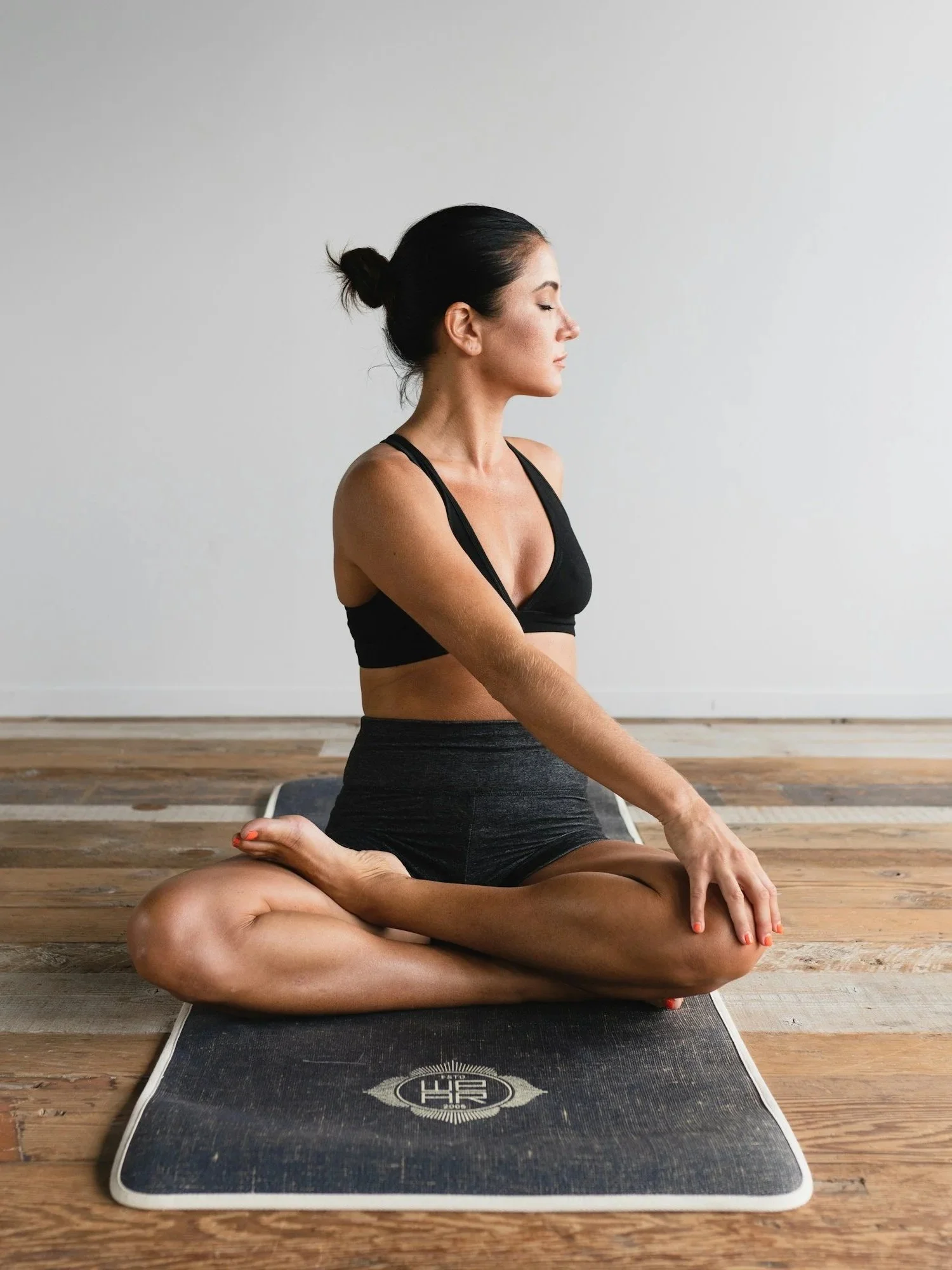 A woman practicing yoga on a black mat in a room with wooden floors and white walls, sitting in a lotus position with her eyes closed.