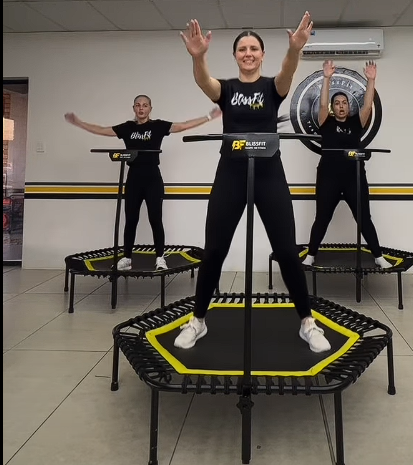 Three women exercising on mini trampolines in a fitness studio, with one woman in front smiling and raising her arms, and two women in the background with arms outstretched.