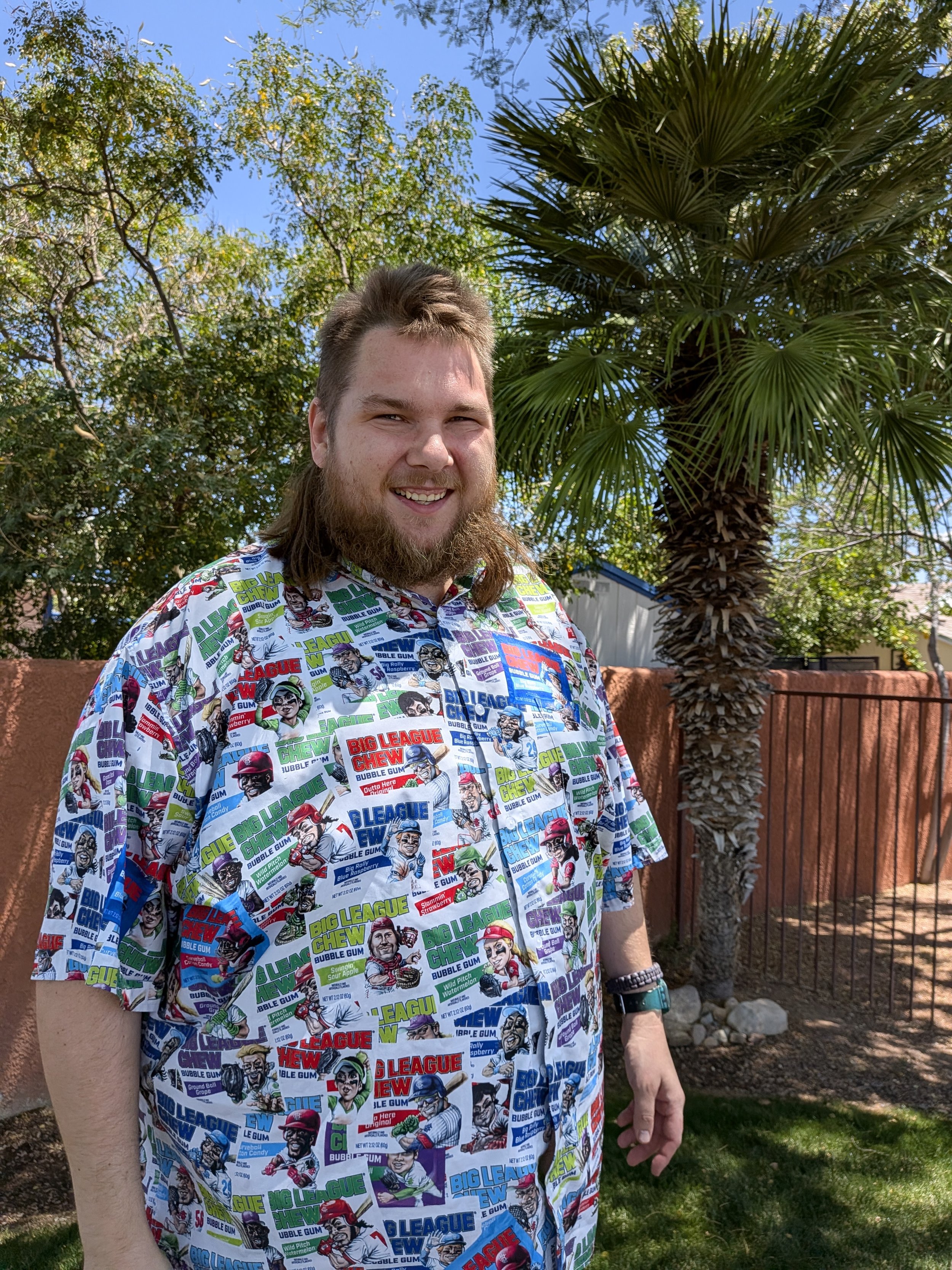 A man with a beard and long hair, smiling, wearing a colorful comic book-themed shirt, standing outdoors next to a tall palm tree and a brick wall during daytime.