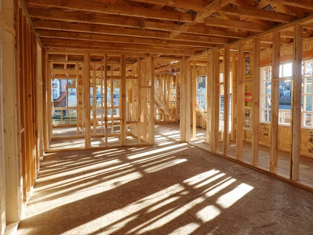 Interior view of a house under construction, showing wooden framing, studs, and exposed ceiling beams. sunlight casts shadows through window openings.