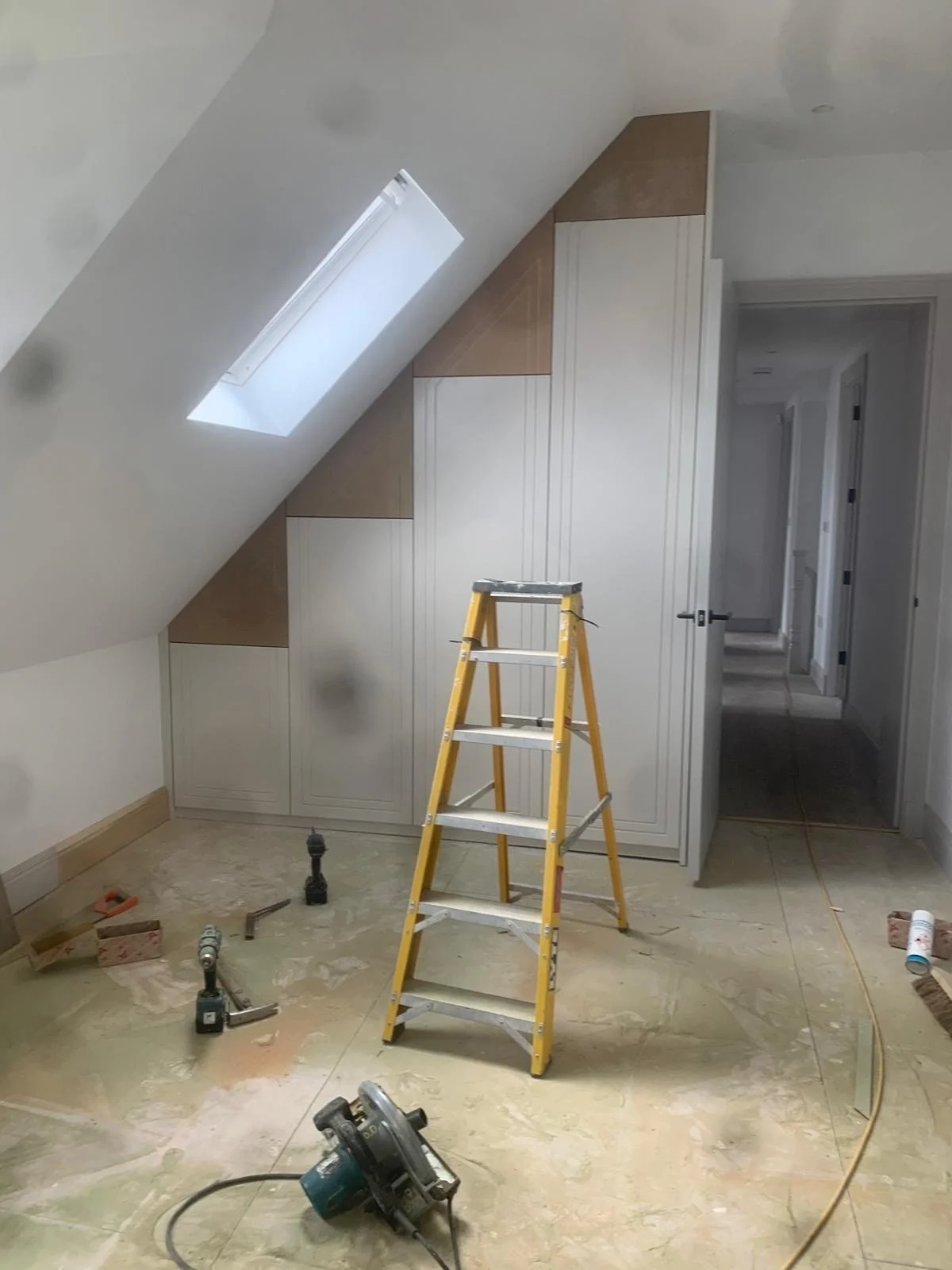 A room under renovation with a yellow ladder in the center, construction tools on the floor, and built-in cabinets with a sloped ceiling and skylight.