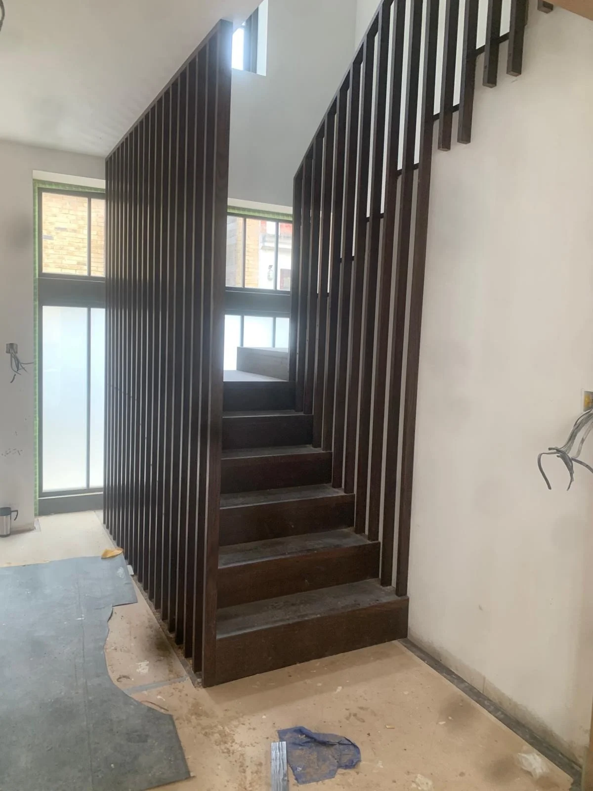 Dark wooden staircase under construction with vertical slats forming a railing, located in a bright room with large windows.