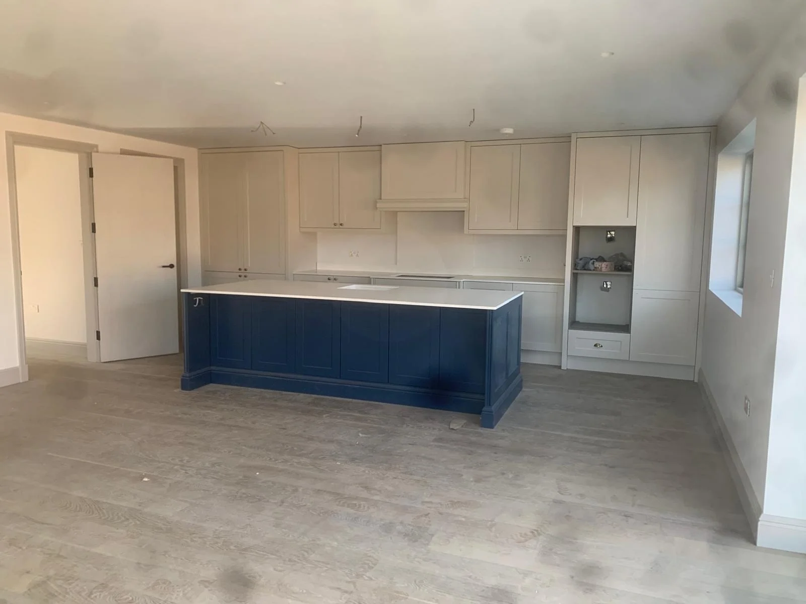 Empty kitchen with light gray cabinets and a blue kitchen island, light wood flooring, and a window on the right wall.