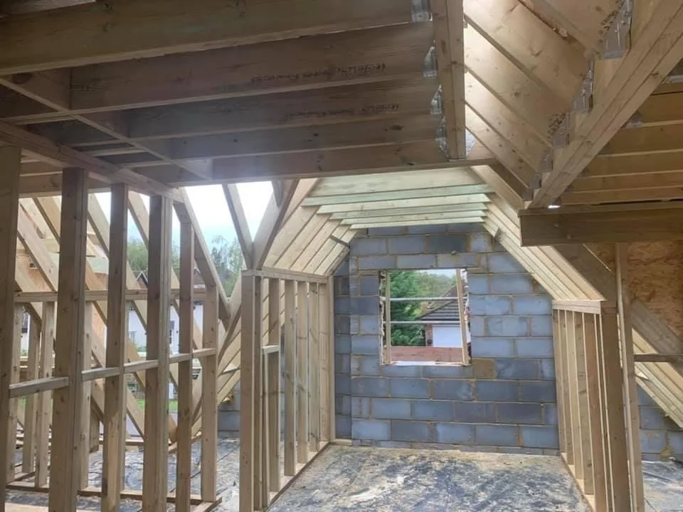Interior view of a house under construction with wooden framing and an incomplete brick wall with a window opening.